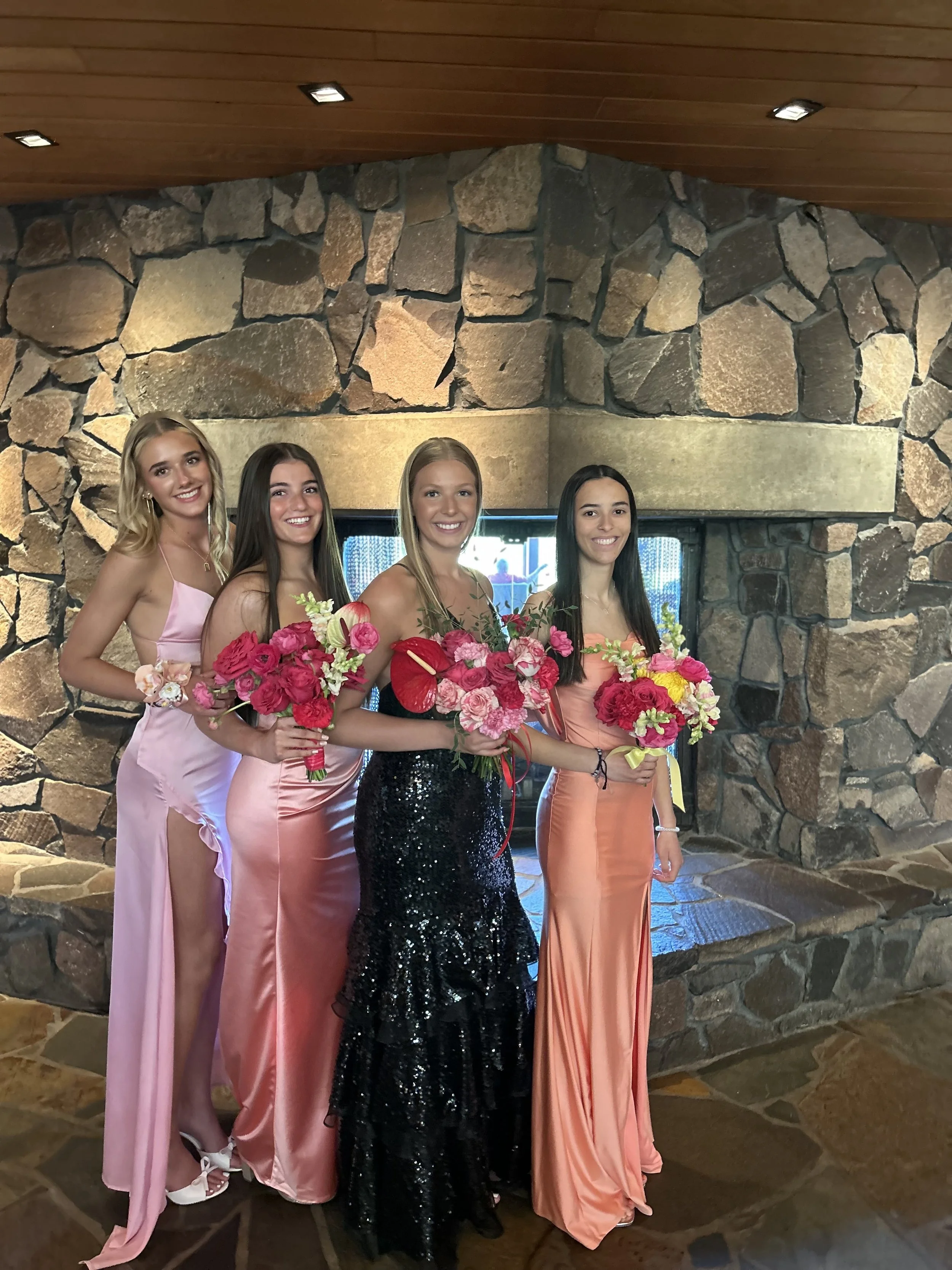 Four young women in elegant dresses holding bouquets of flowers, standing in front of a stone fireplace.
