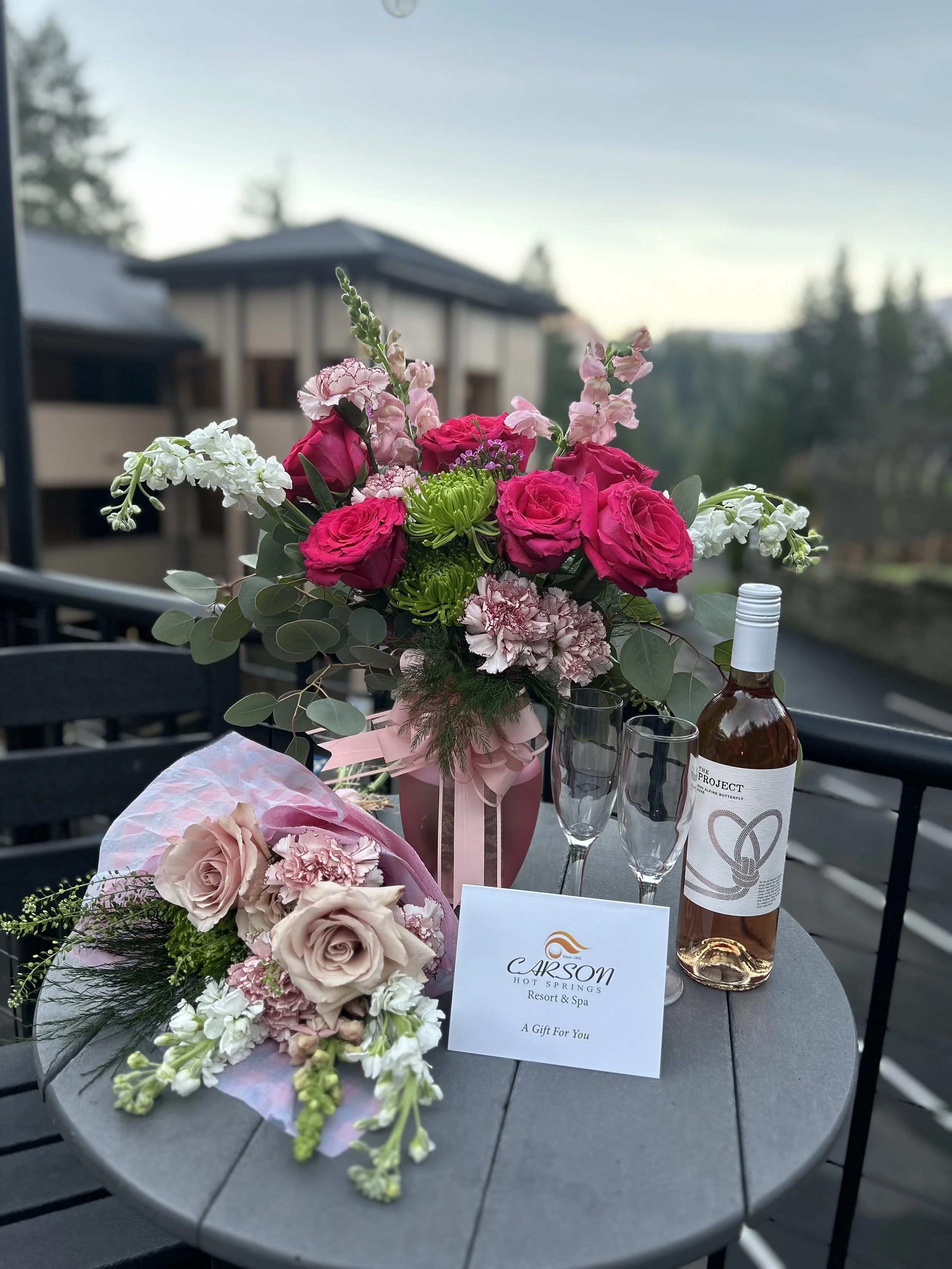 A table with a flower bouquet, a pink gift box with flowers, a card from Carson Hot Springs Resort & Spa, a bottle of rosé wine, and two empty champagne glasses on a balcony at sunset.