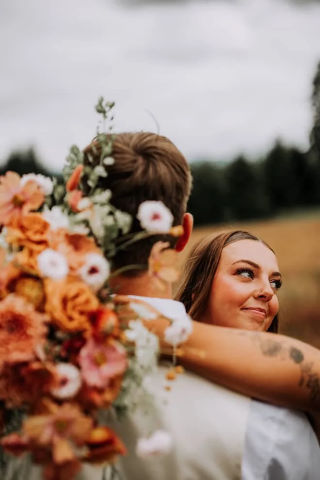 A woman lovingly hugging a man holding a large bouquet of colorful flowers outdoors on a cloudy day.