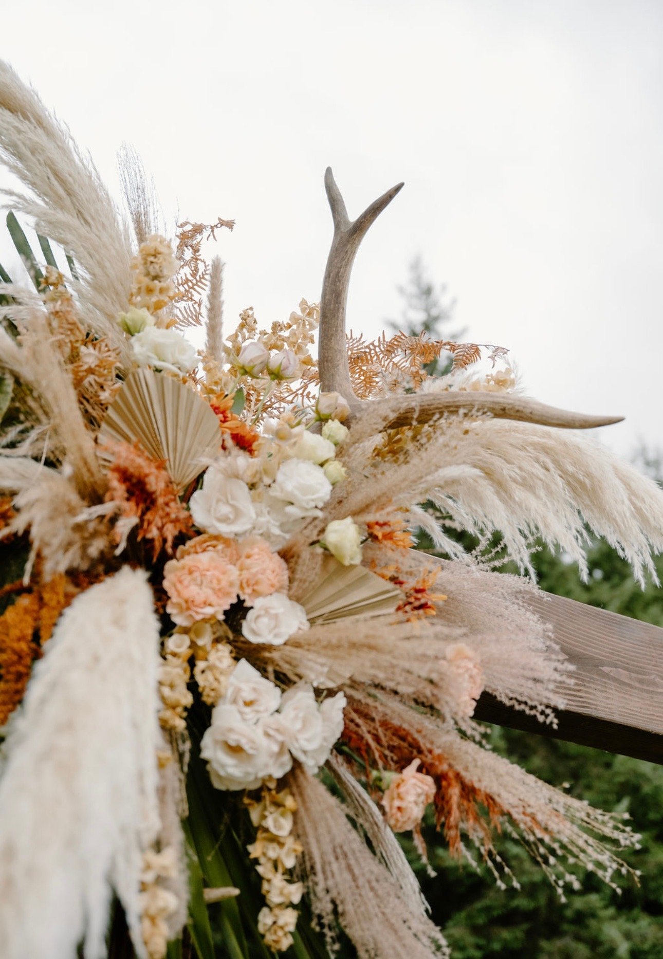 Decorative floral arrangement featuring white and peach flowers, dried grasses, burlap, and a small antler piece, set outdoors against a cloudy sky.