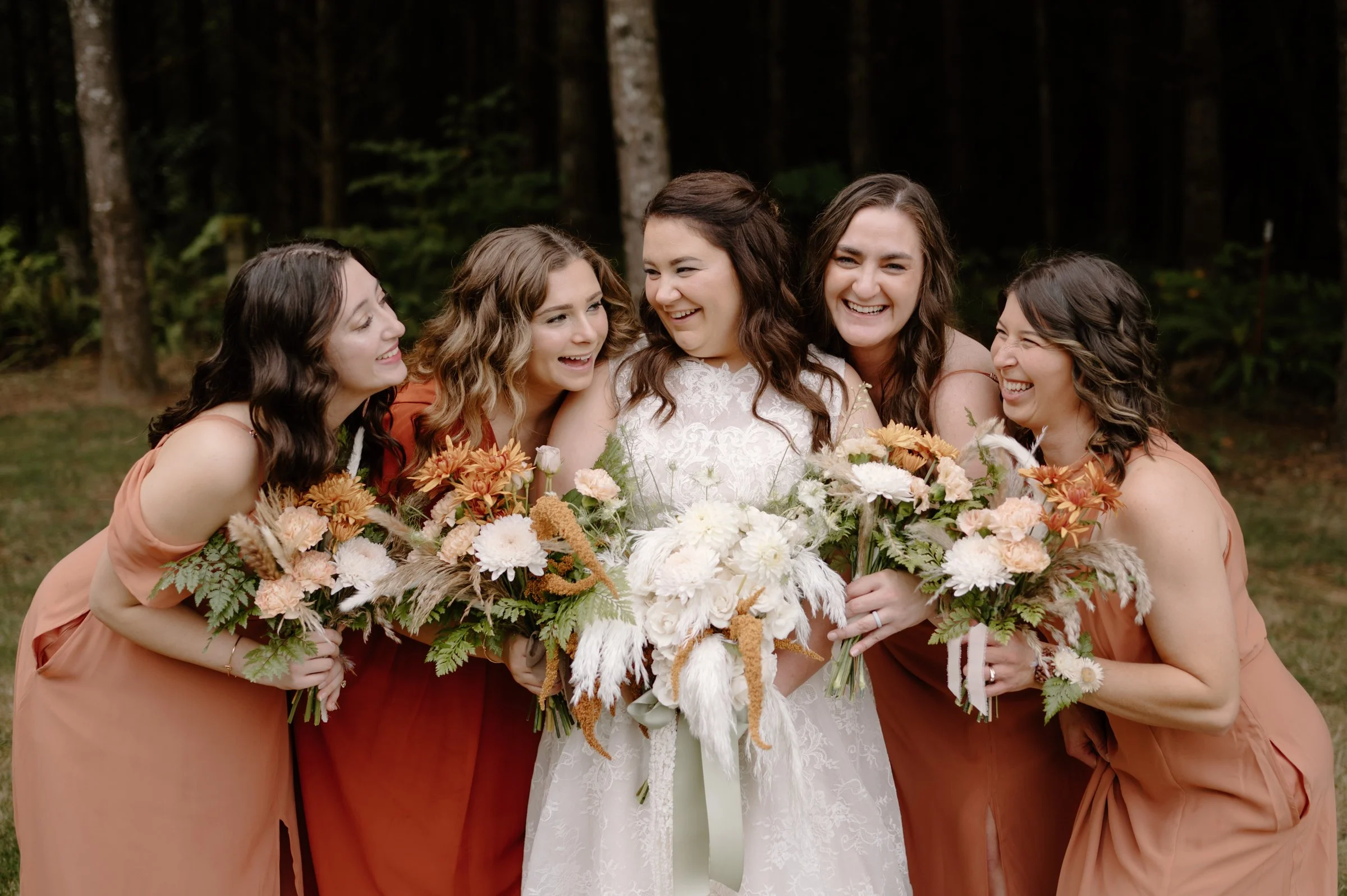A bride in a white lace wedding dress holding a bouquet of white flowers, surrounded by five women in peach-colored dresses holding bouquets of mixed flowers, standing outdoors in a wooded area, all smiling and laughing together.
