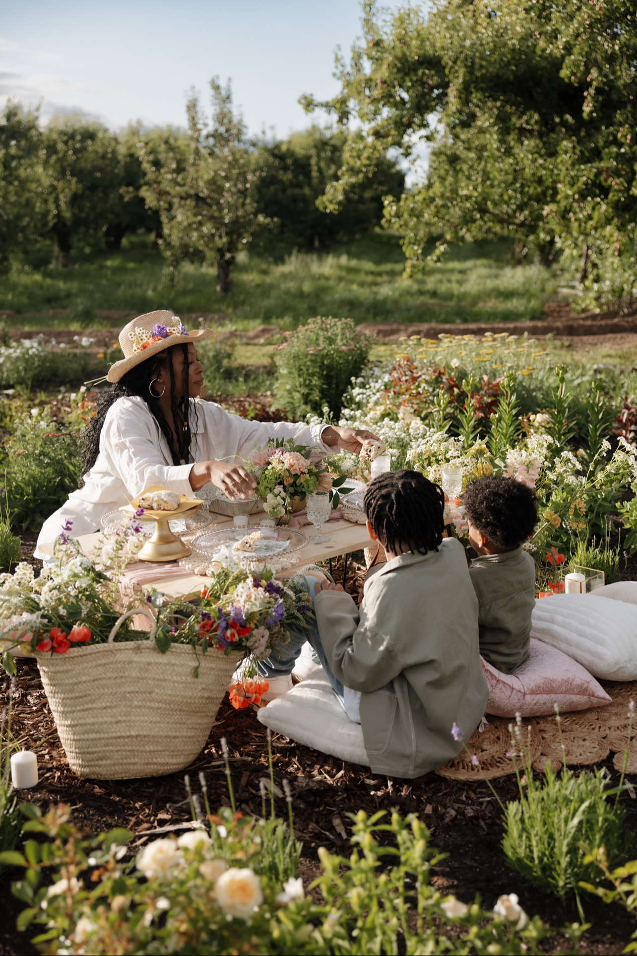 A woman and two children having a picnic in a garden surrounded by flowers and trees. The woman is wearing a straw hat and white shirt, and they are seated on cushions around a low table with flowers and food.