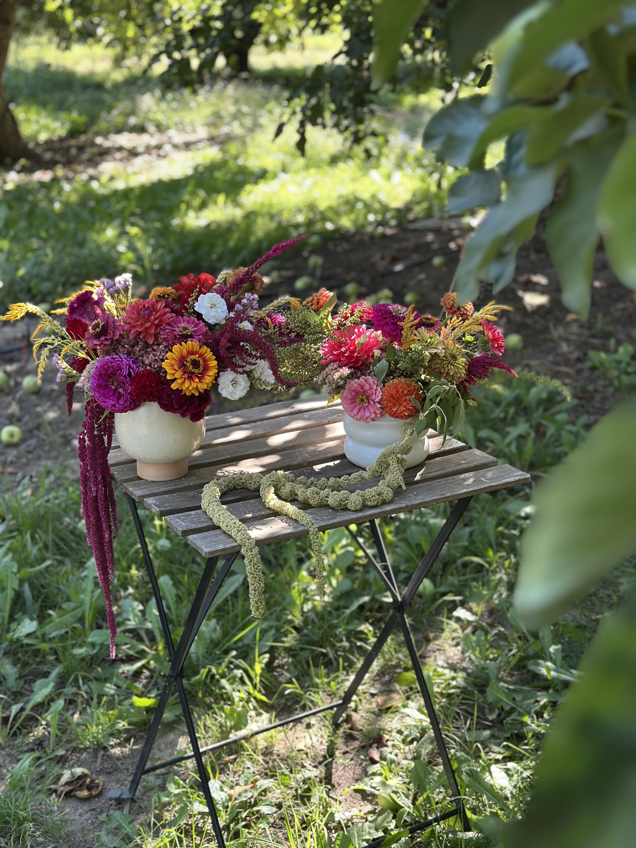 Two white vases with colorful flowers placed on a small wooden table outdoors in a garden setting, with greenery and trees in the background.