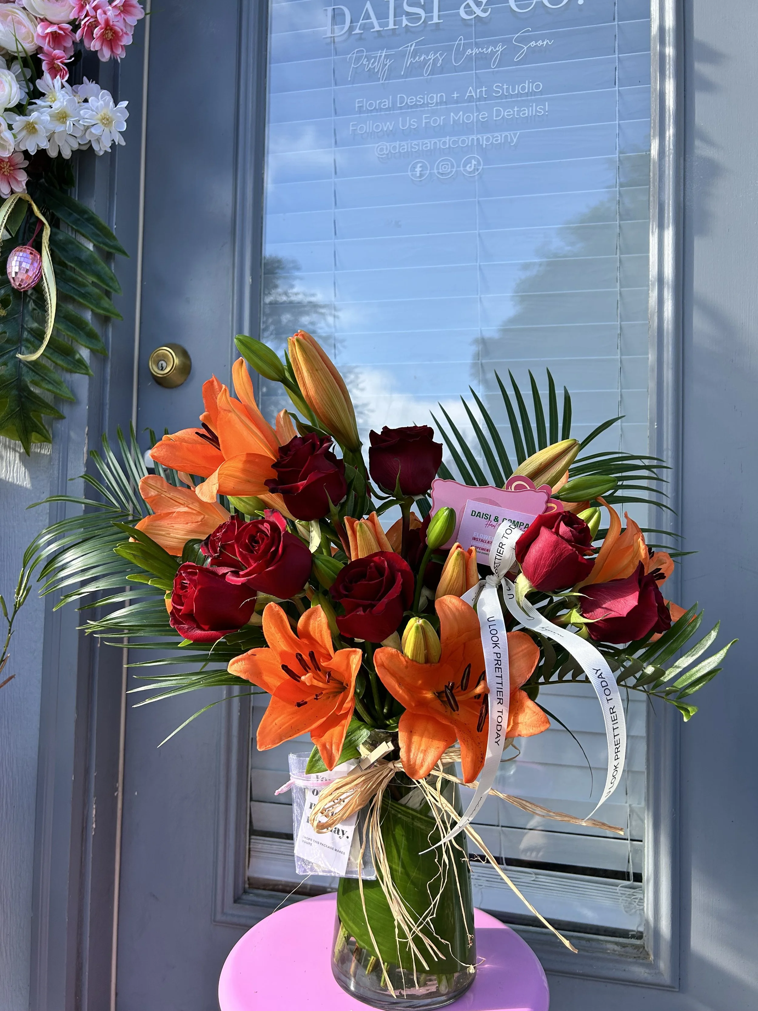 A bouquet of red roses and orange lilies in a glass vase on a pink table outside a floral design studio.