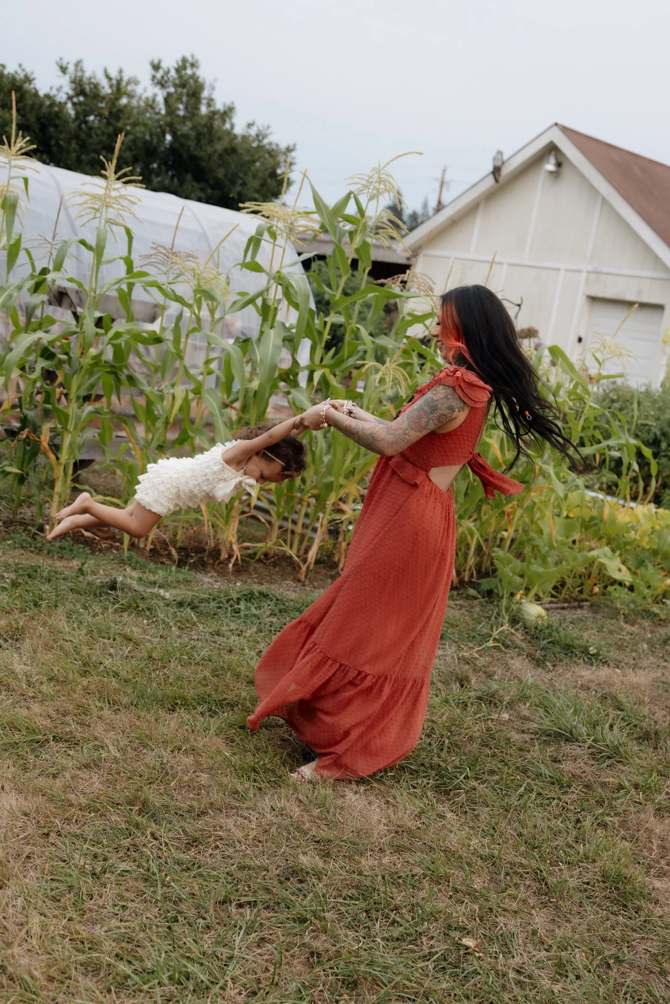 A woman is twirling a young girl holding hands outdoors near a garden of corn plants; the woman has long black hair, tattoos, and is wearing a long rust-colored dress. The girl is in a white dress, and they are smiling and enjoying a playful moment.