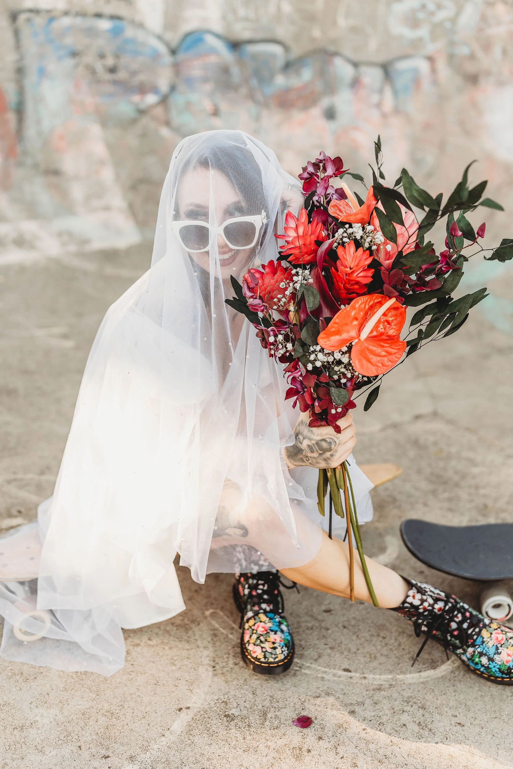 Person dressed as a bride with sunglasses and a veil, sitting on the ground, holding a large bouquet of vibrant red and orange flowers, wearing colorful floral boots, with a skateboard nearby, against a graffiti-covered wall backdrop.