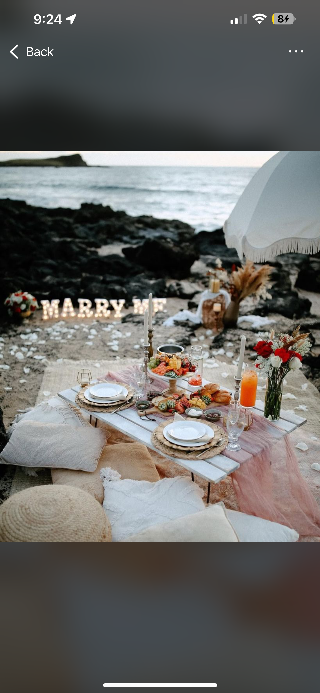 Beachside picnic setup with a low table, plates, candles, flowers, and a decorative "Marry Me" sign near the ocean