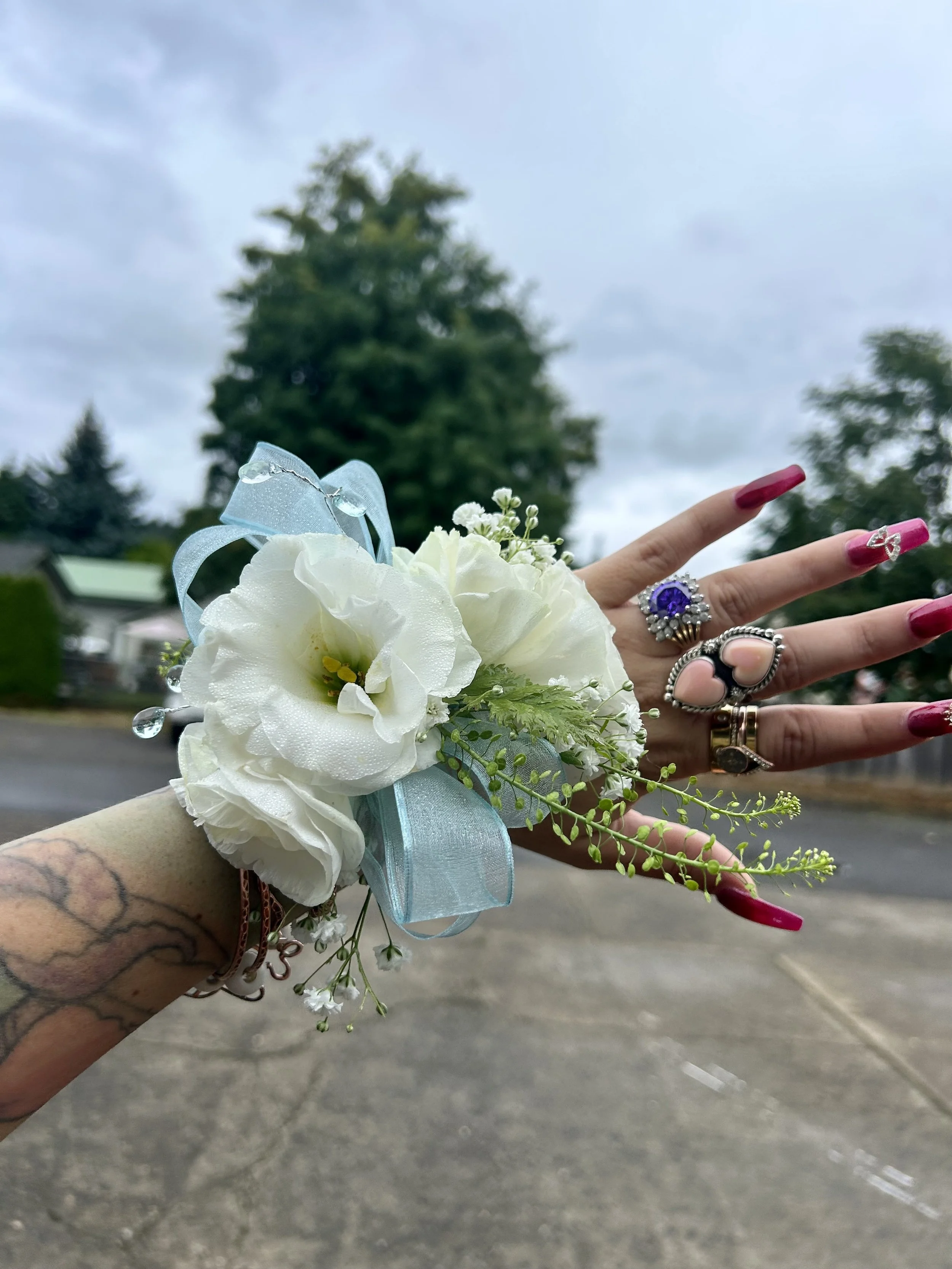 A person's arm with rings and long pink nails extended, holding a boutonniere with white flowers, greenery, and a light blue ribbon, outdoors on a cloudy day.