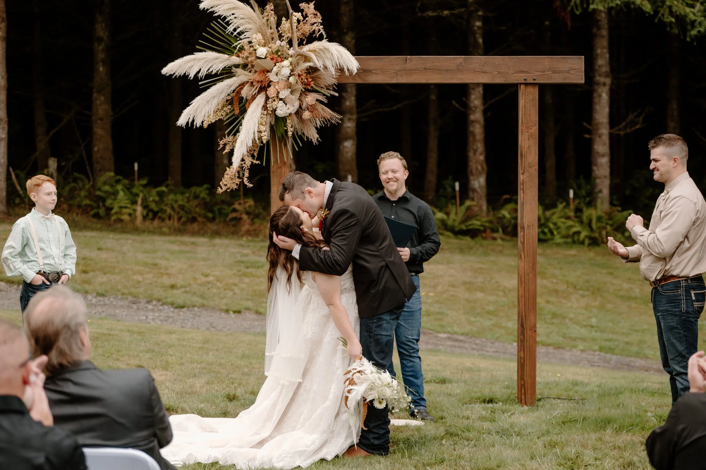 A couple is kissing at an outdoor wedding ceremony beneath a wooden arch decorated with floral arrangements. The bride wears a white wedding dress, and the groom wears a dark suit. Guests and officiants are visible, with a forest background.
