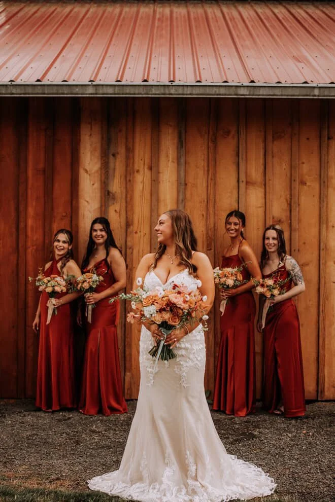 A bride in a white wedding gown holding a large bouquet of flowers, surrounded by four bridesmaids in red dresses holding smaller bouquets, standing in front of a wooden barn wall.