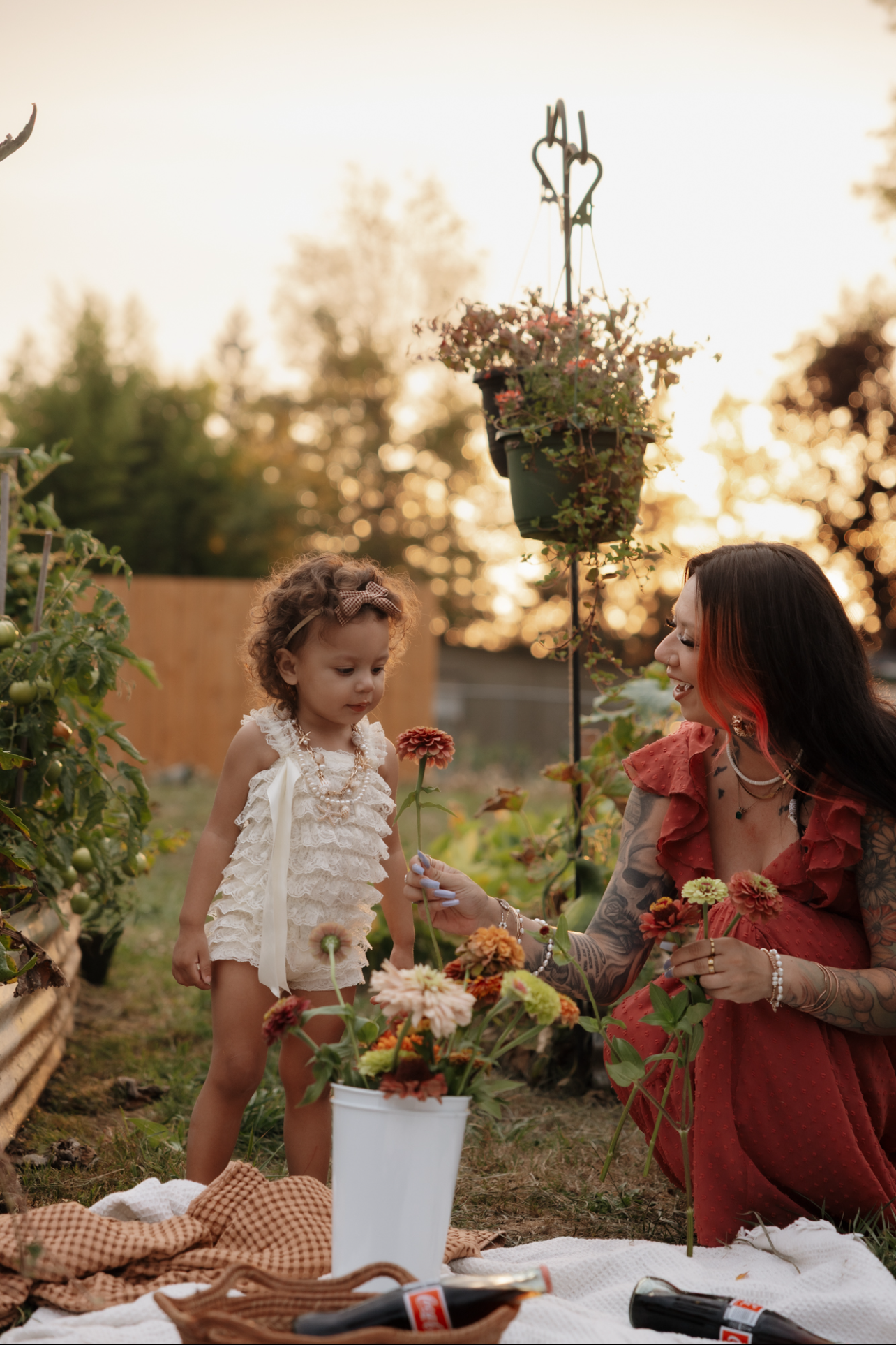 A woman with tattoos and bright red hair kneels on a white blanket, smiling at a young girl in a cream-colored dress with a bow in her curly hair. The woman presents a flower to the girl, surrounded by a garden with plants and colorful flowers during sunset.