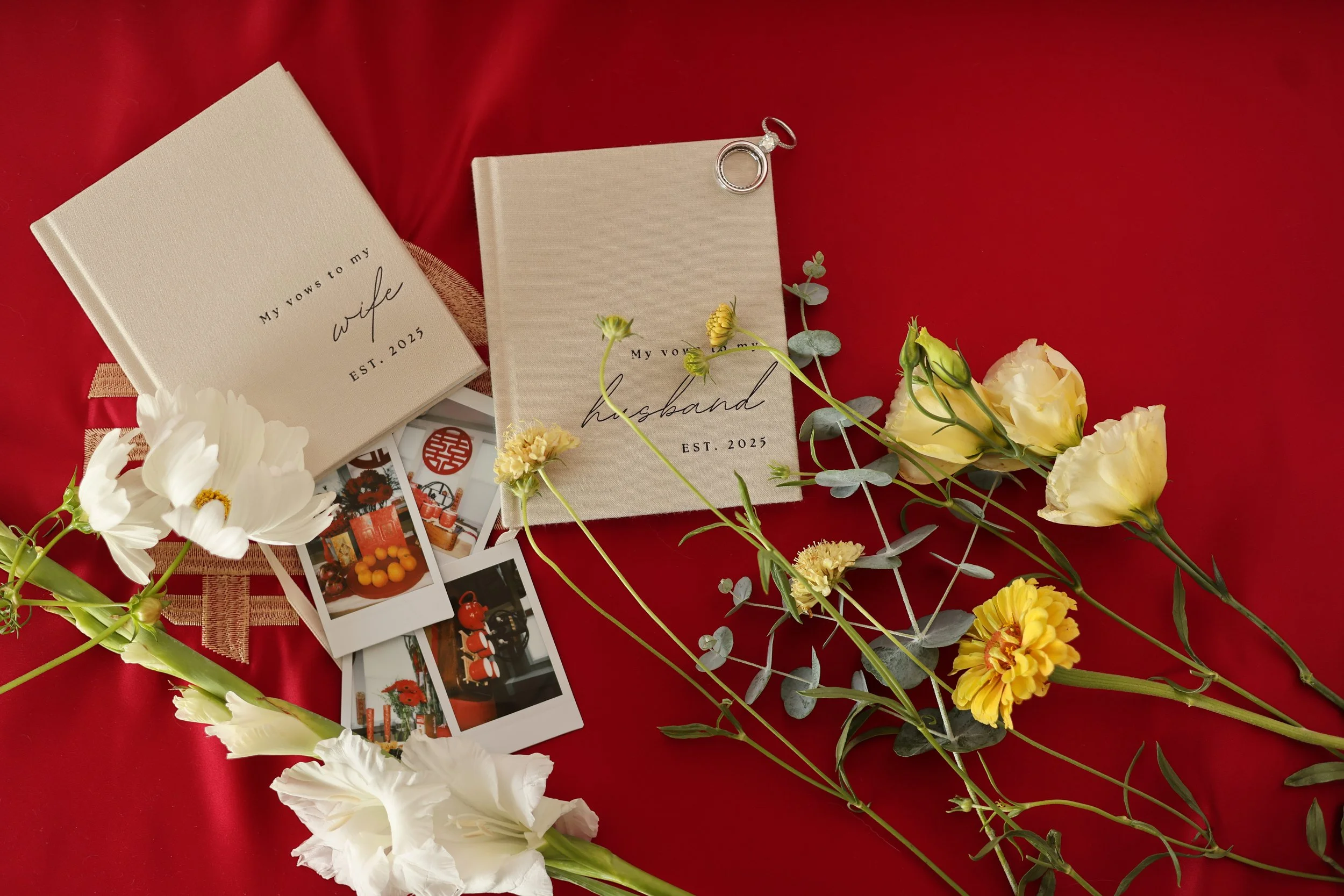 Wedding vow books labeled "My vows to my wife" and "My vows to my husband" on a red satin cloth, surrounded by white and yellow flowers and several instant photographs.