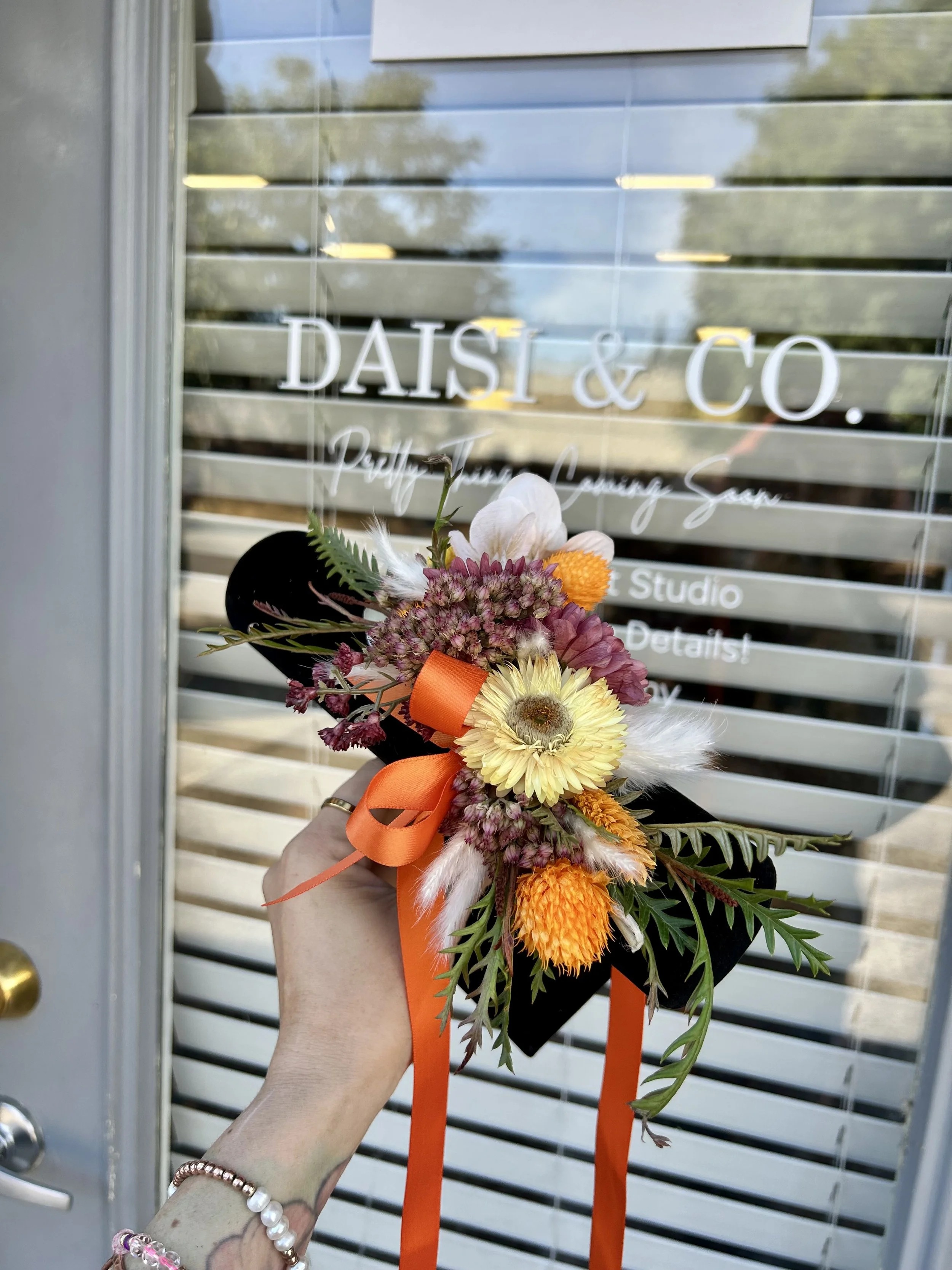 Person holding a colorful bouquet of flowers with orange ribbons in front of a glass door with the text 'Daisy & Co.' written on it.