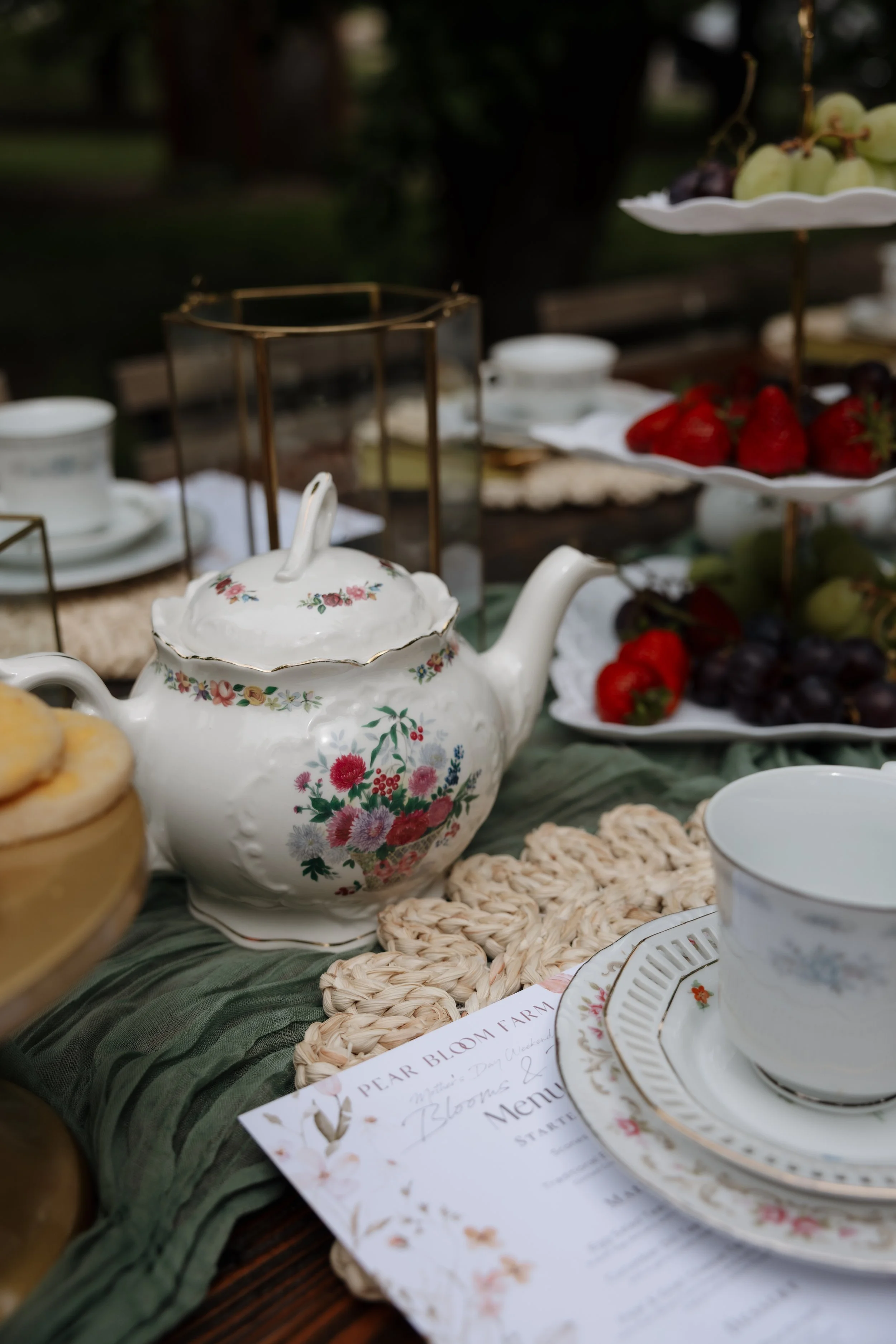 A vintage floral teapot surrounded by a fruit display with strawberries, grapes, and a tiered stand, along with teacups, plates, and a menu on a vintage table setting for an outdoor tea party.