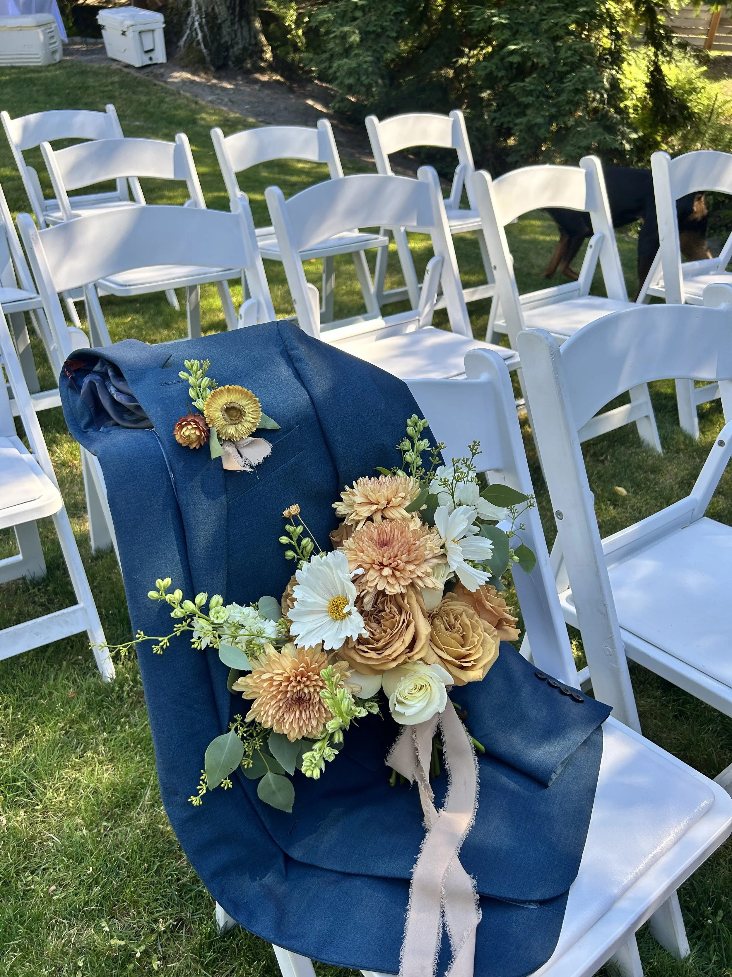 A blue suit jacket with a floral boutonniere rests on a white chair, surrounded by white chairs arranged outdoors, with trees and grass in the background.