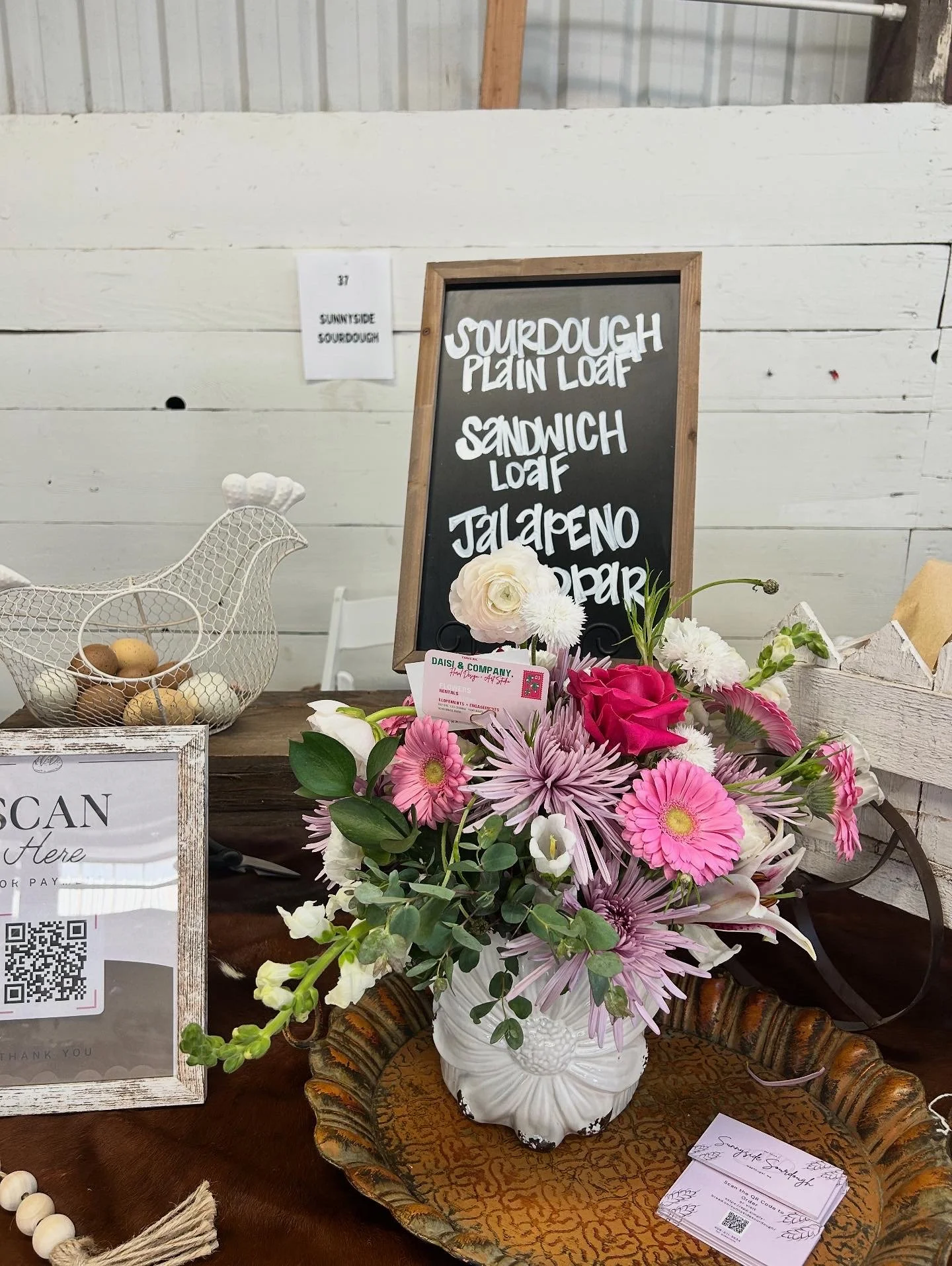 A bouquet of pink and white flowers in a white vase on a decorative tray, with a chalkboard menu listing sourdough plain loaf, sandwich loaf, jalapeño, and bagel. There is also a small framed sign with a QR code and a wire basket with eggs in the background.