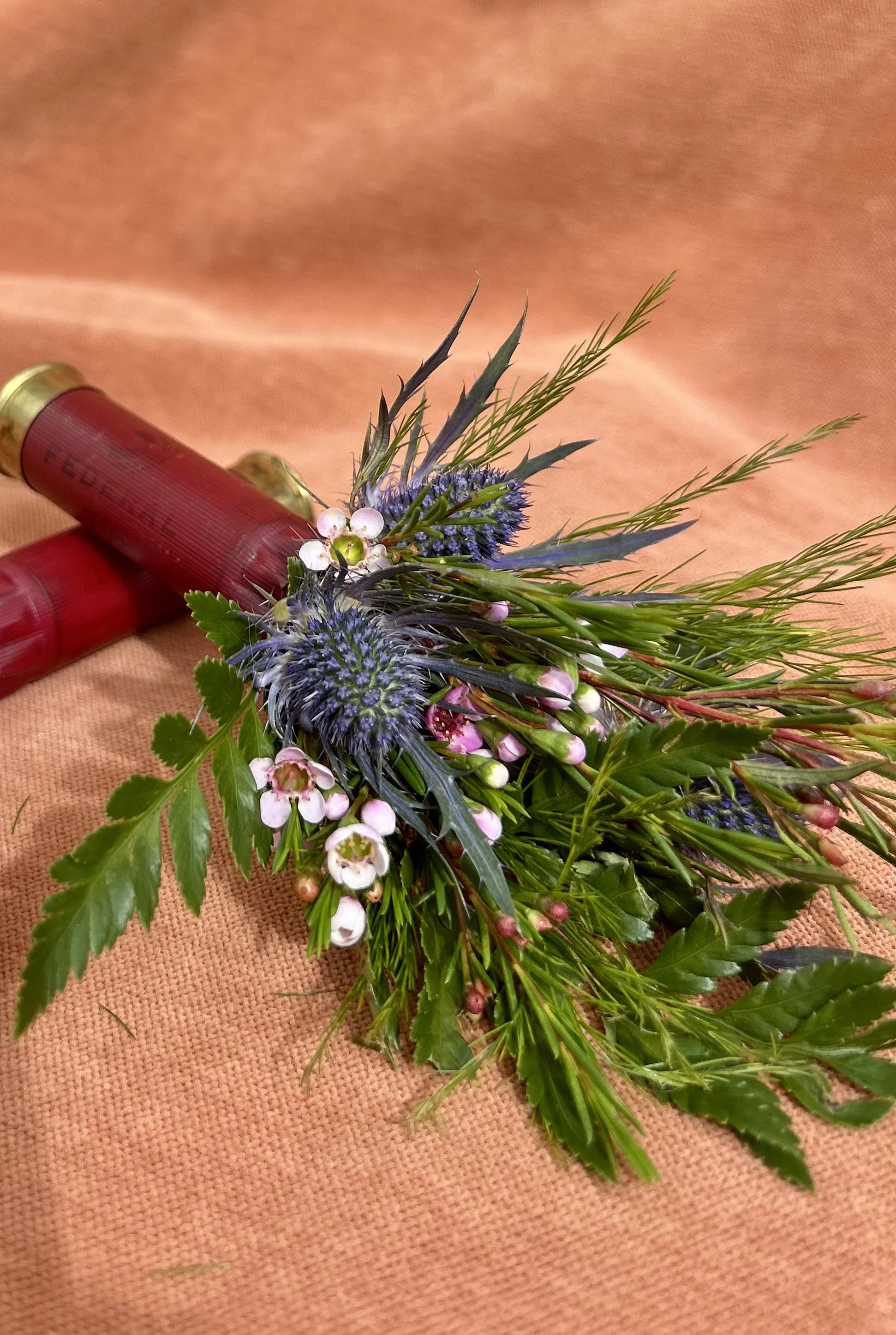 A bouquet of wildflowers with purple thistles, small white and pink flowers, and greenery, placed beside two red shotgun shells on a peach-colored fabric background.