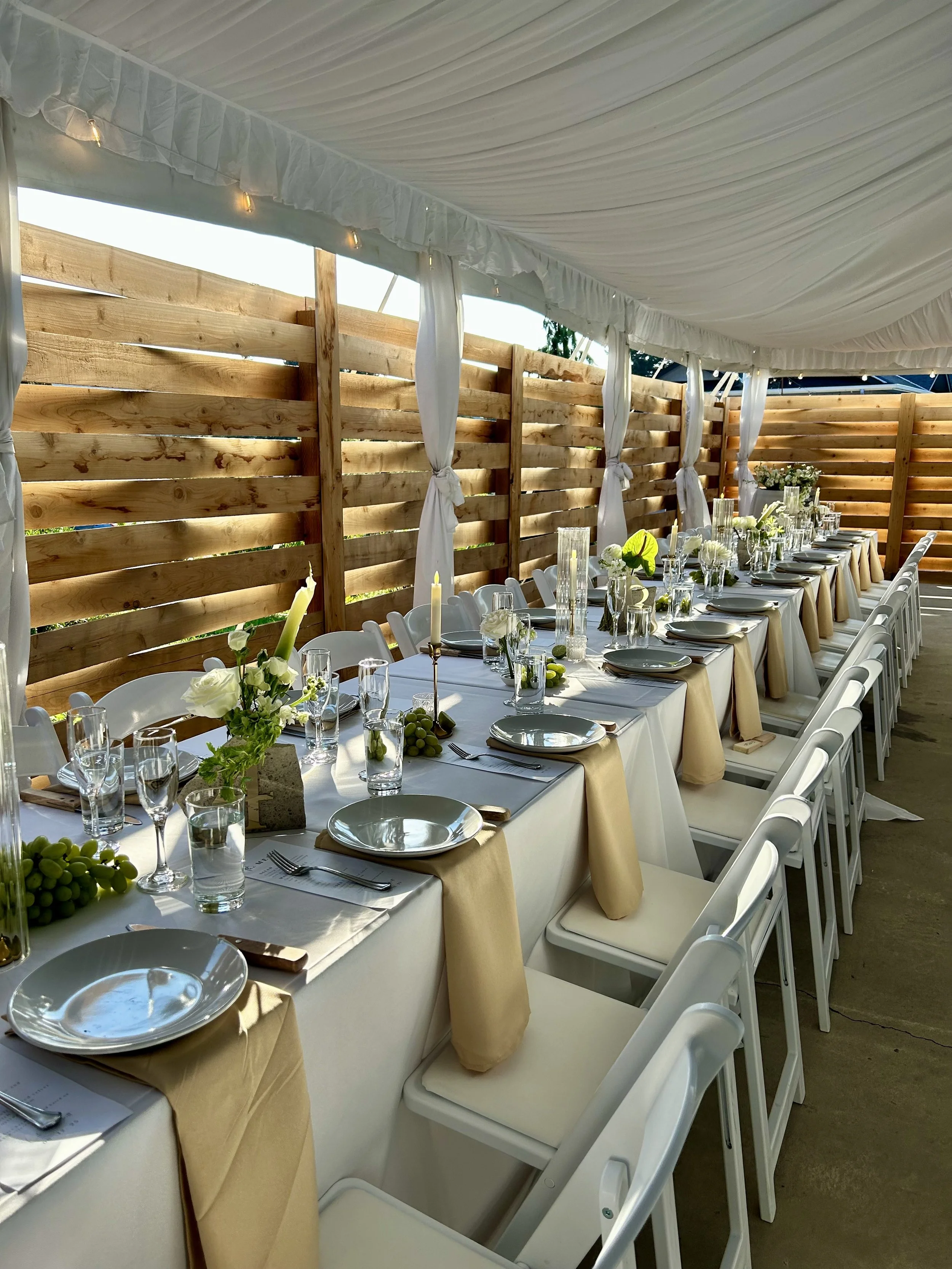 A long banquet table set for a celebration, with white tablecloths, beige napkins, plates, glasses, and centerpieces of flowers and candles, under a white canopy with string lights and a wooden fence in the background.