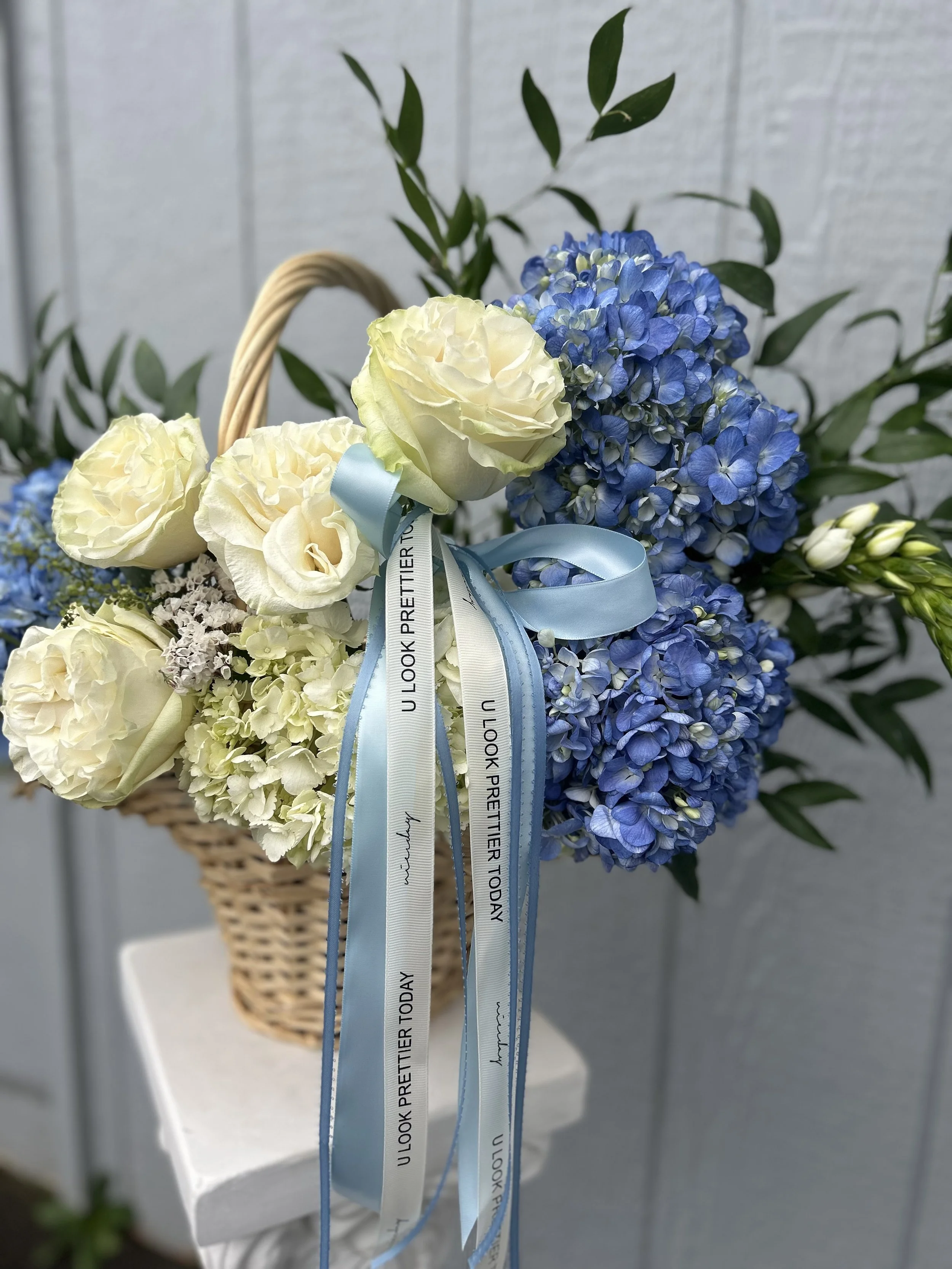 A flower arrangement in a wicker basket with blue hydrangeas, white roses, and other white flowers, decorated with blue and white ribbons that say 'U LOOK PRETTIER TODAY'.