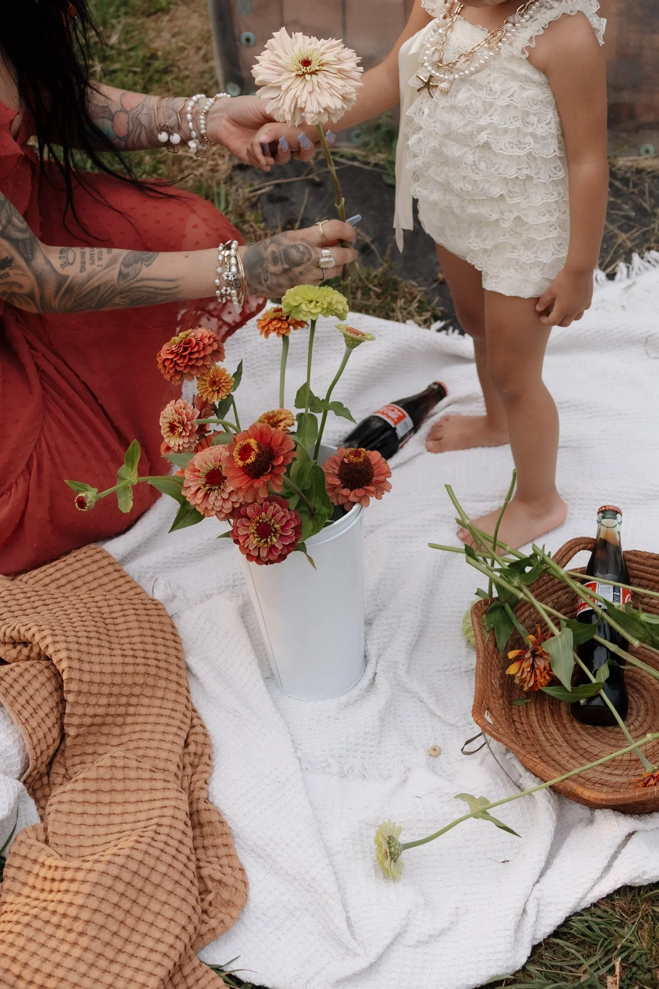 A woman with tattooed arms and jewelry hands a pink flower to a young girl in a white lace dress. They are on a white blanket surrounded by a bouquet of colorful flowers, a basket, and beer bottles, outdoors.