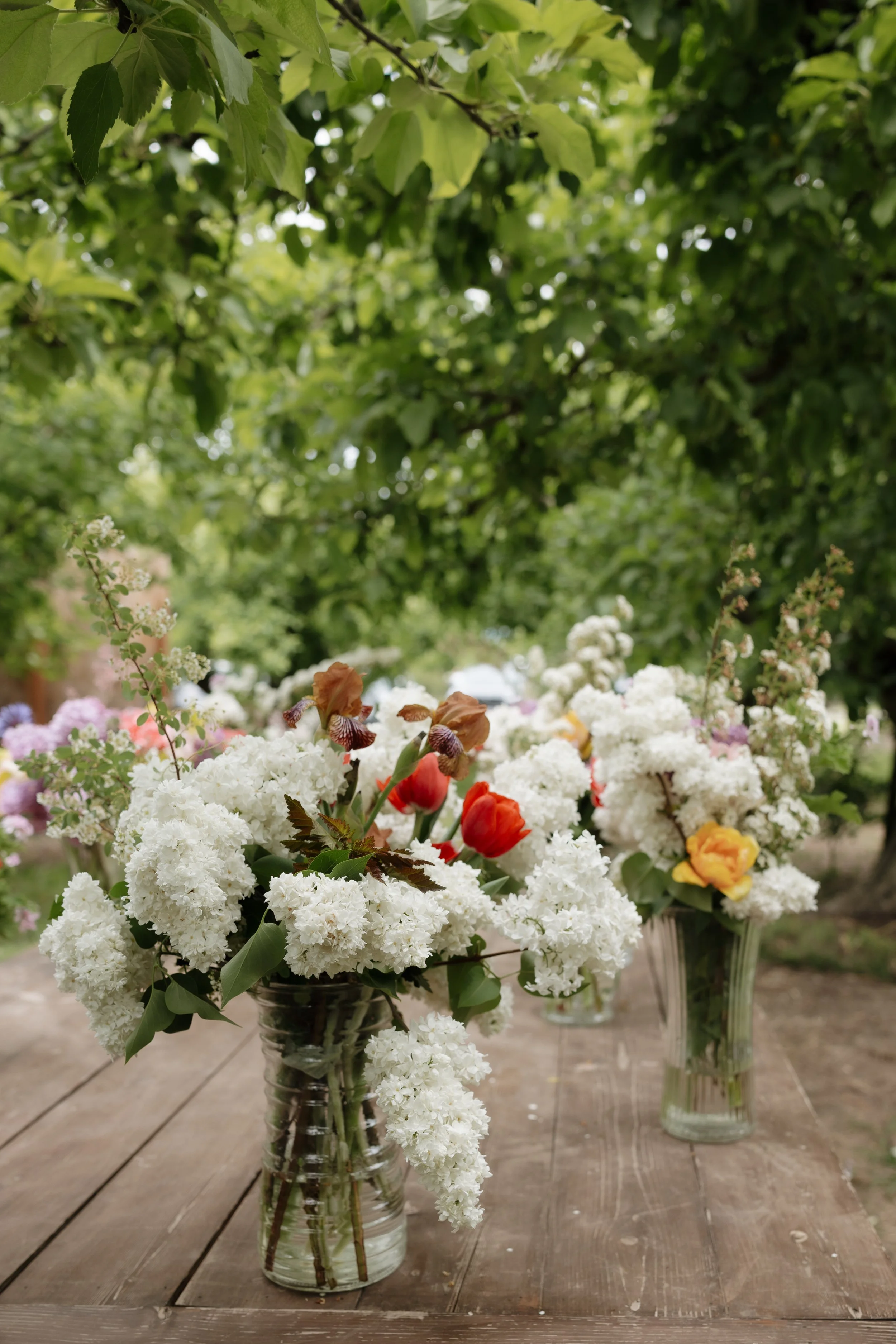 Three glass vases filled with spring flowers, including tulips, hyacinths, and other blossoms, arranged on a wooden table outdoors beneath green leafy trees.