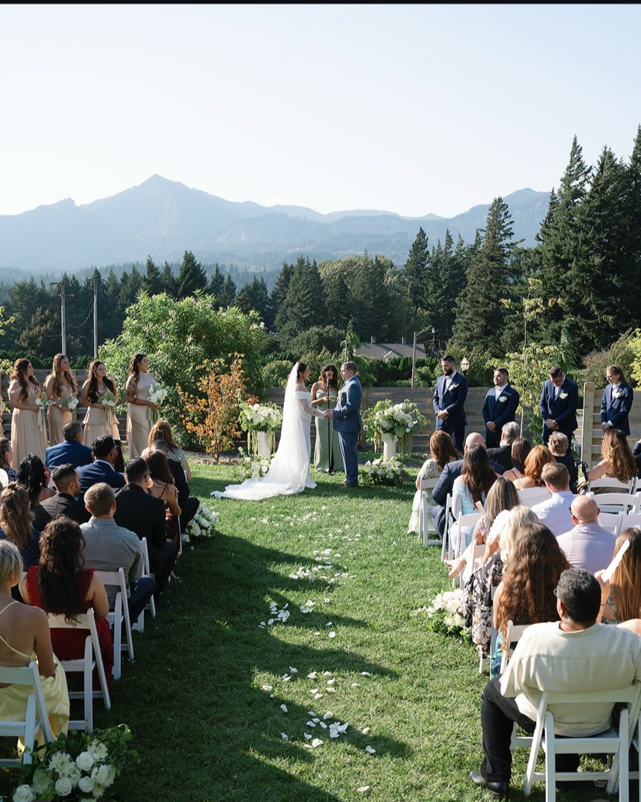 An outdoor wedding ceremony on a grassy lawn with a mountain backdrop, featuring a bride and groom exchanging vows, surrounded by bridesmaids and groomsmen, with seated guests watching.