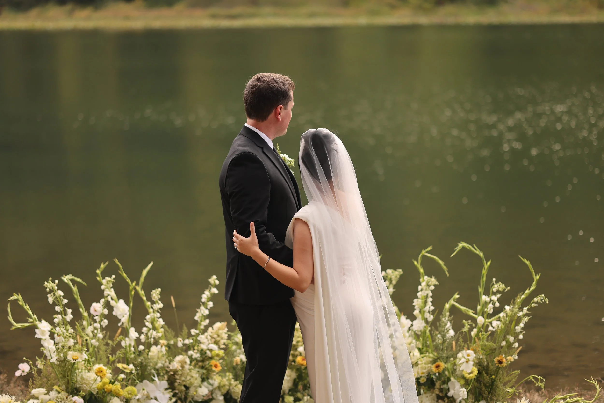 A bride and groom stand by a lake surrounded by flowers, facing each other during their wedding ceremony.