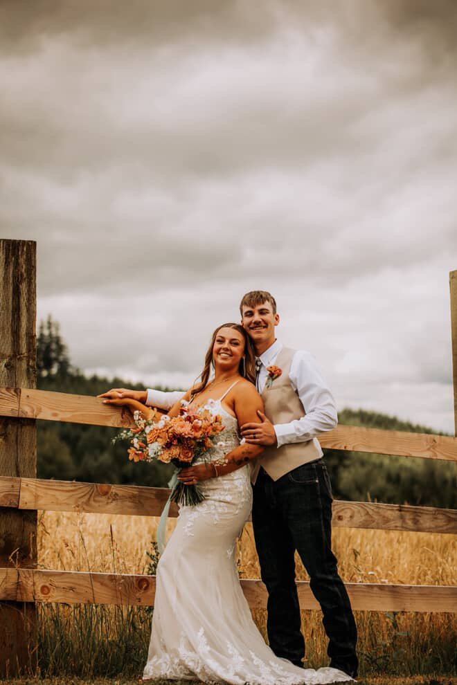A bride and groom stand together outdoors in front of a wooden fence, smiling and embracing. The bride holds a bouquet of flowers, wearing a white wedding dress, and the groom is dressed in a white shirt, light vest, and dark pants. The background shows a cloudy sky and a natural landscape.