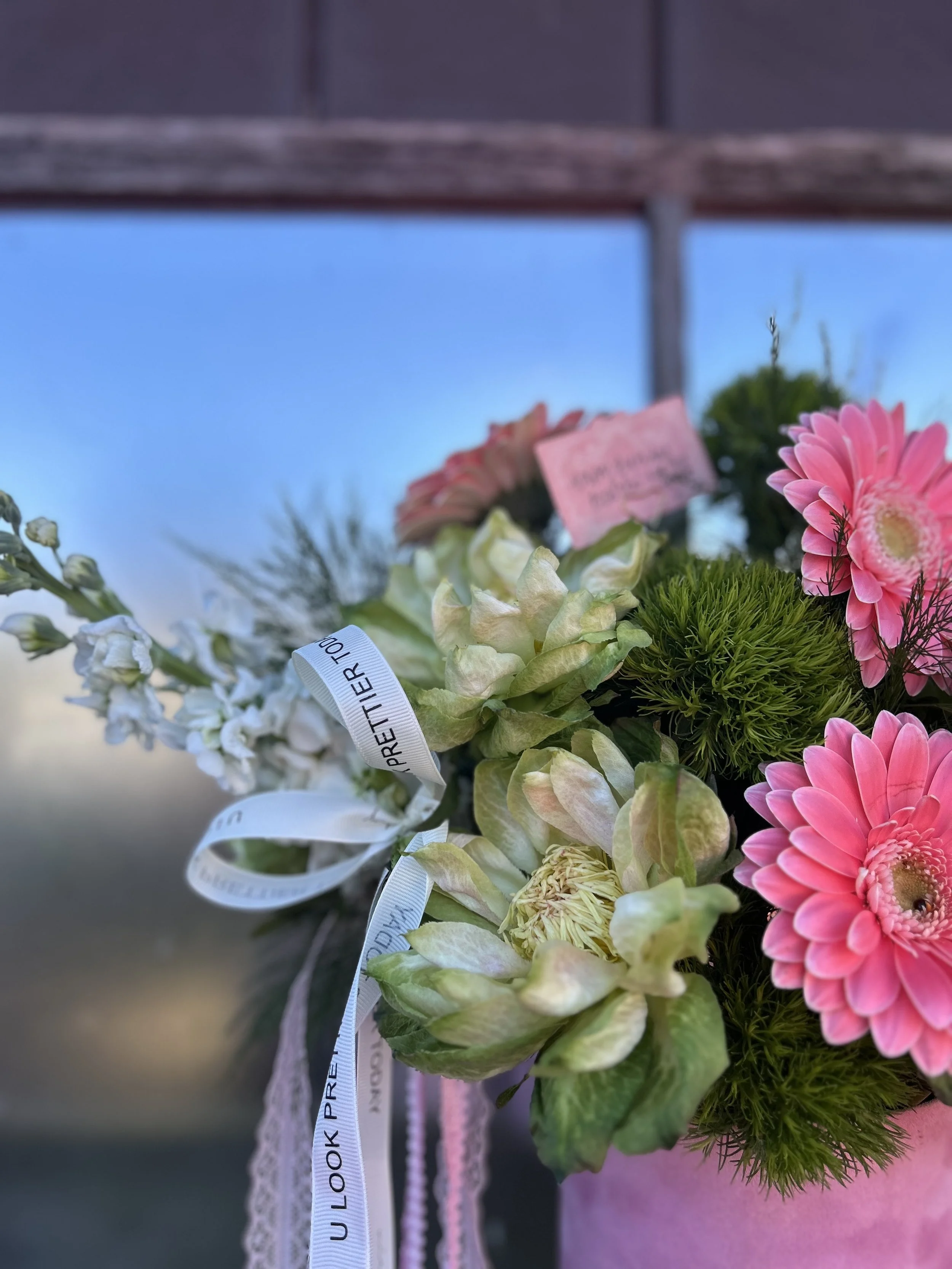 Close-up of a bouquet of pink and white flowers with green foliage, tied with white ribbons, against a blurred outdoor background and blue sky.