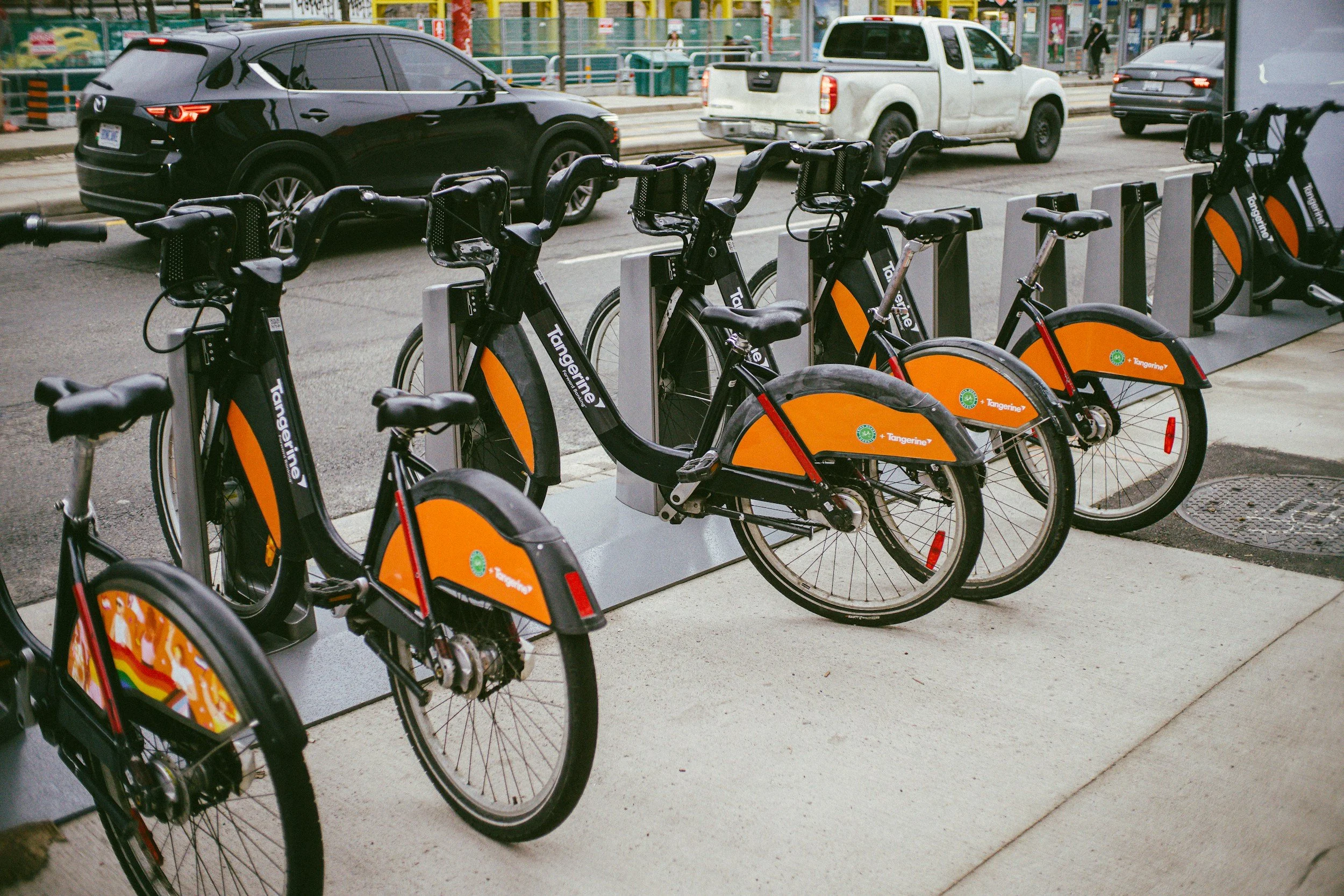 Bike share station with orange and black bicycles parked on the sidewalk. Parked cars on the street are visible in the background.