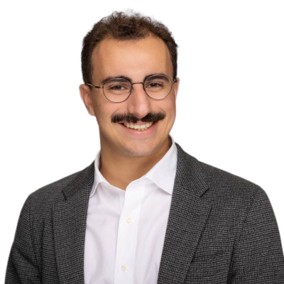 Gabe Blanc with glasses, a mustache, and short curly hair smiling, wearing a white shirt and a dark gray blazer.