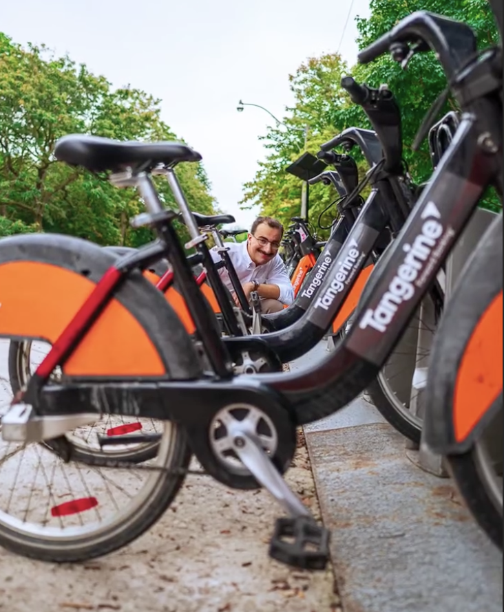 Gabe Blanc while sitting between a row of black and orange Tangarine electric bikes outdoors on a paved path, with green trees and a cloudy sky in the background.