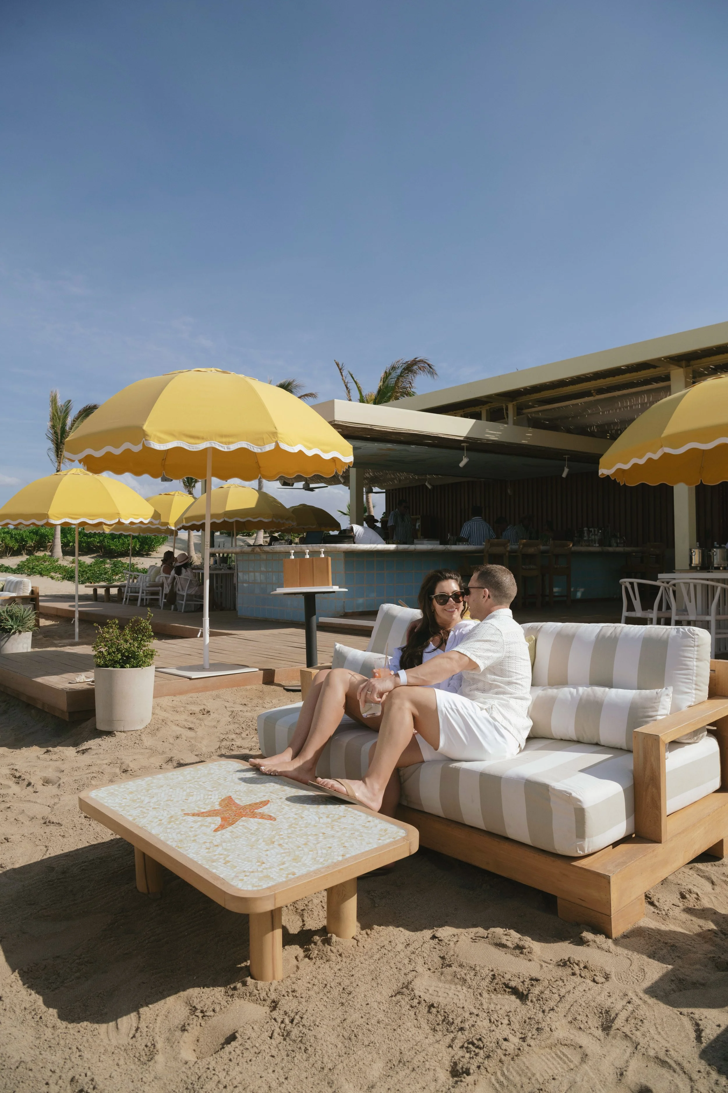 A man and woman sitting on a large outdoor sofa on a beach with sand, under a sunny sky. They are smiling and holding drinks, with yellow umbrellas and a bar in the background.
