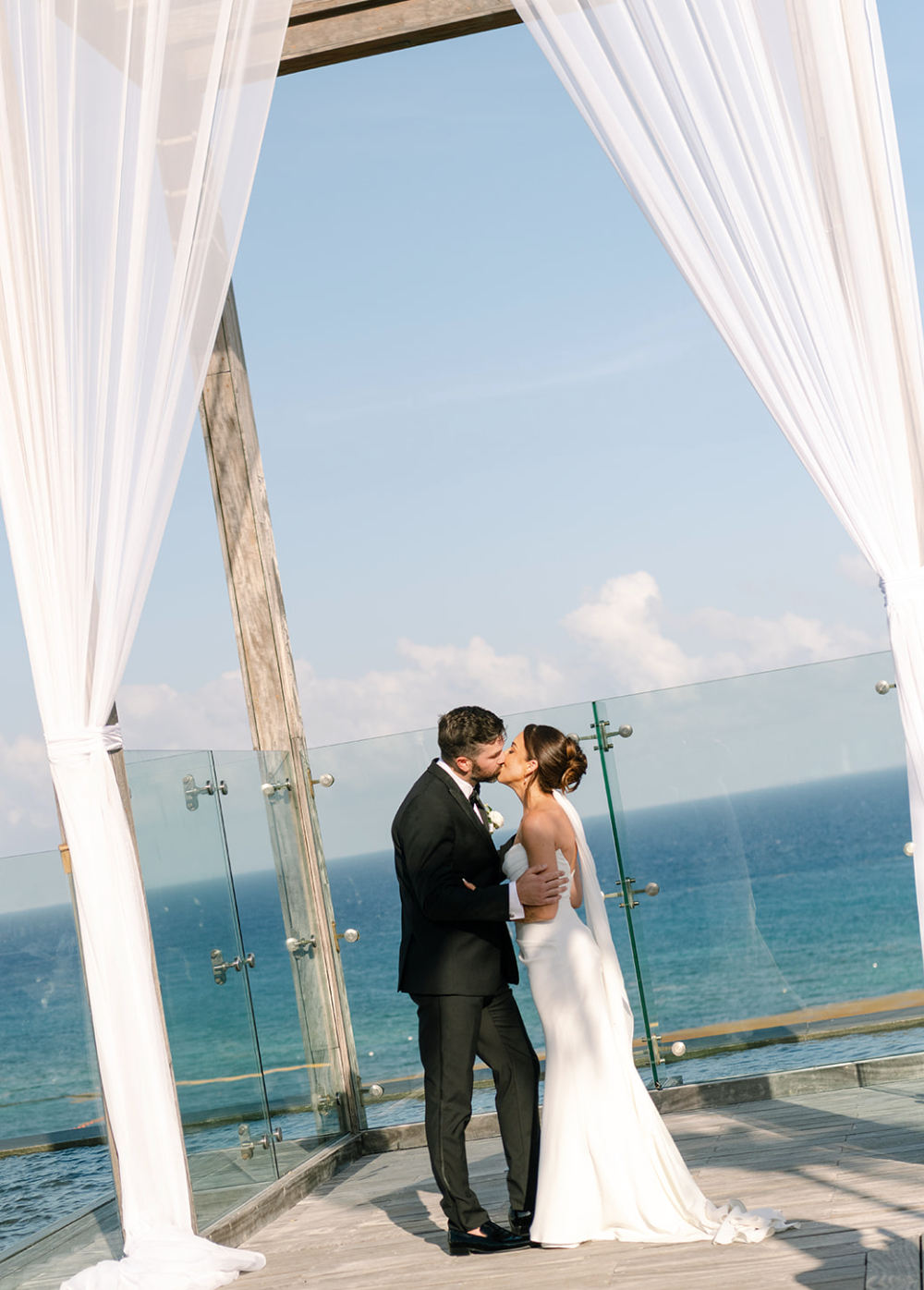 A bride and groom share a kiss on a seaside wedding altar with white curtains and a glass railing, overlooking the ocean and sky.