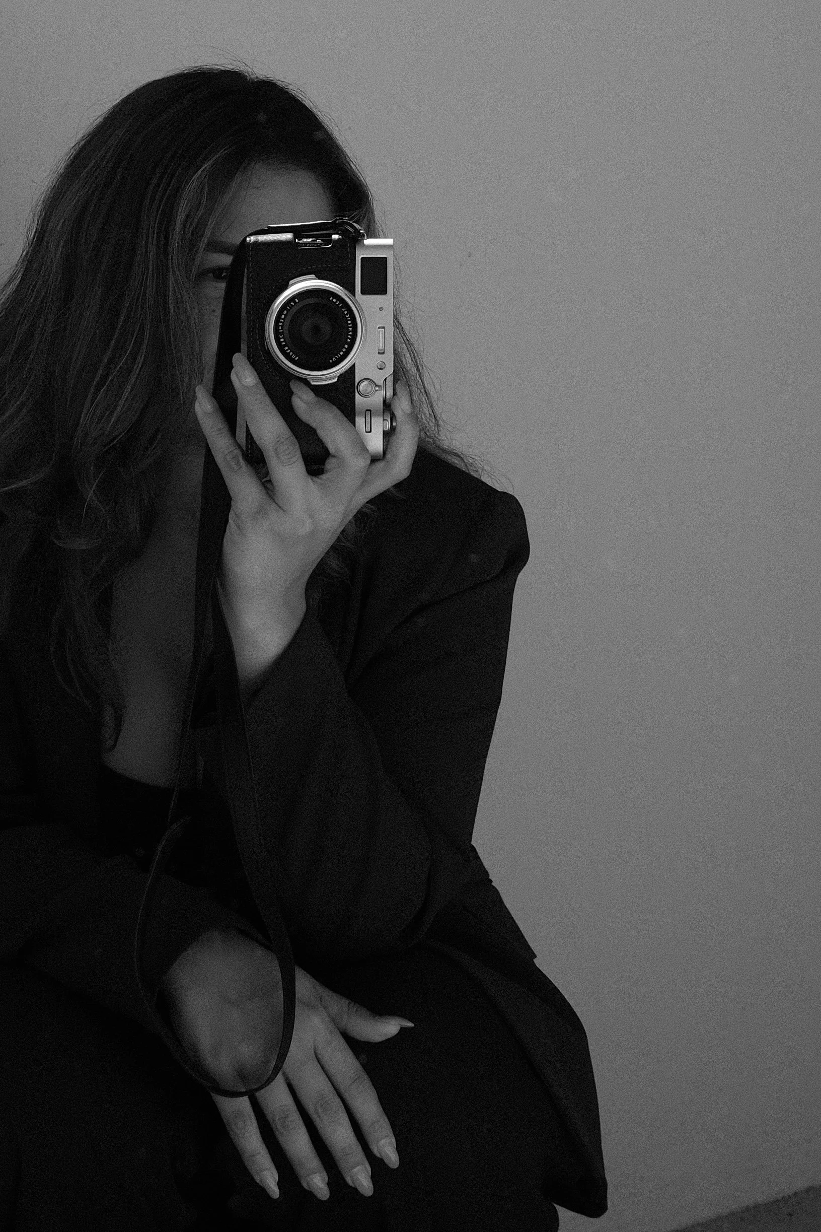 A woman with long wavy hair, partially obscured face, holding a camera up to her eye, taking a self-portrait in black and white.