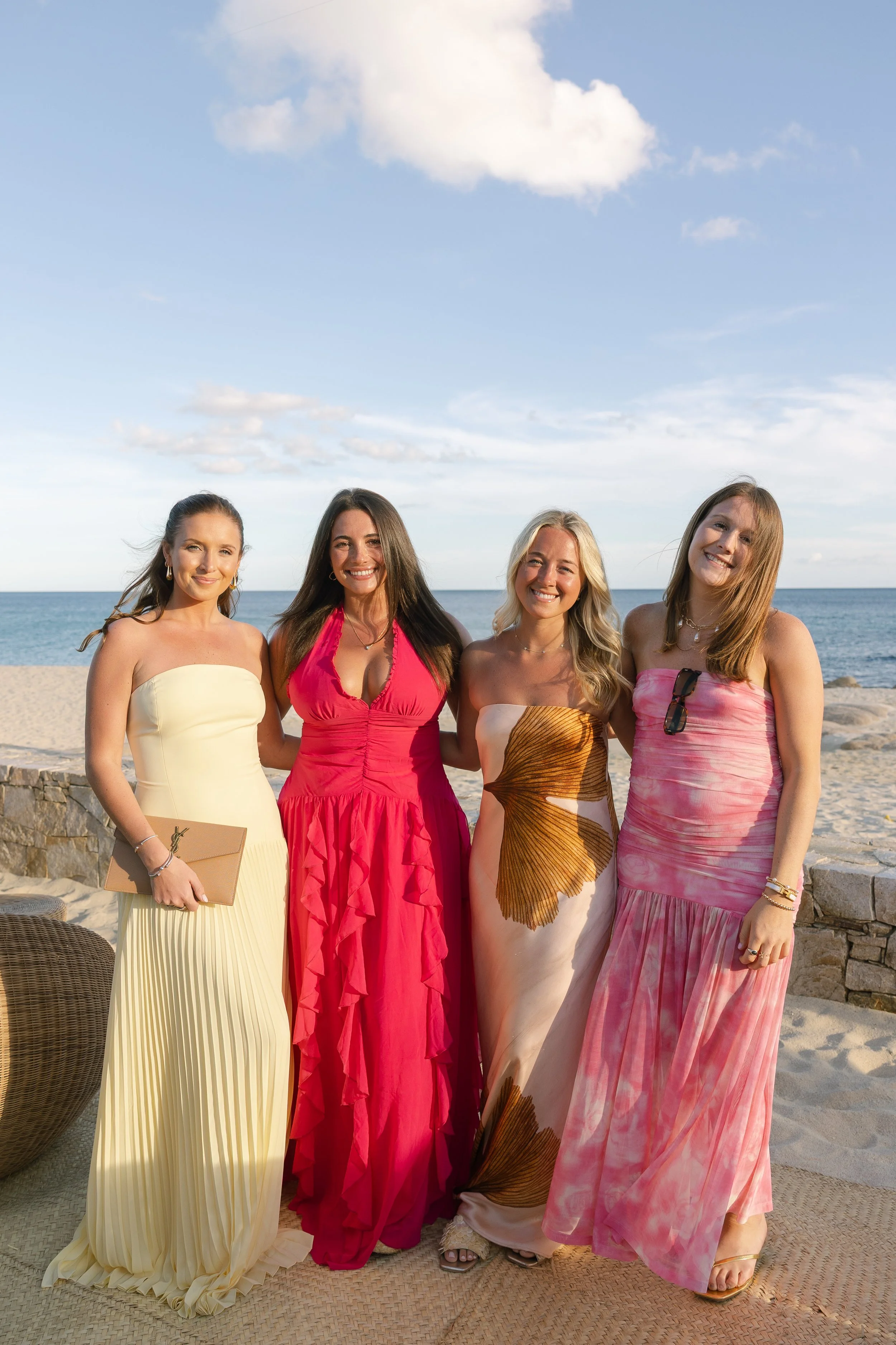 Four women standing on a beach with the ocean and sky in the background, all wearing colorful, elegant dresses and smiling at the camera.