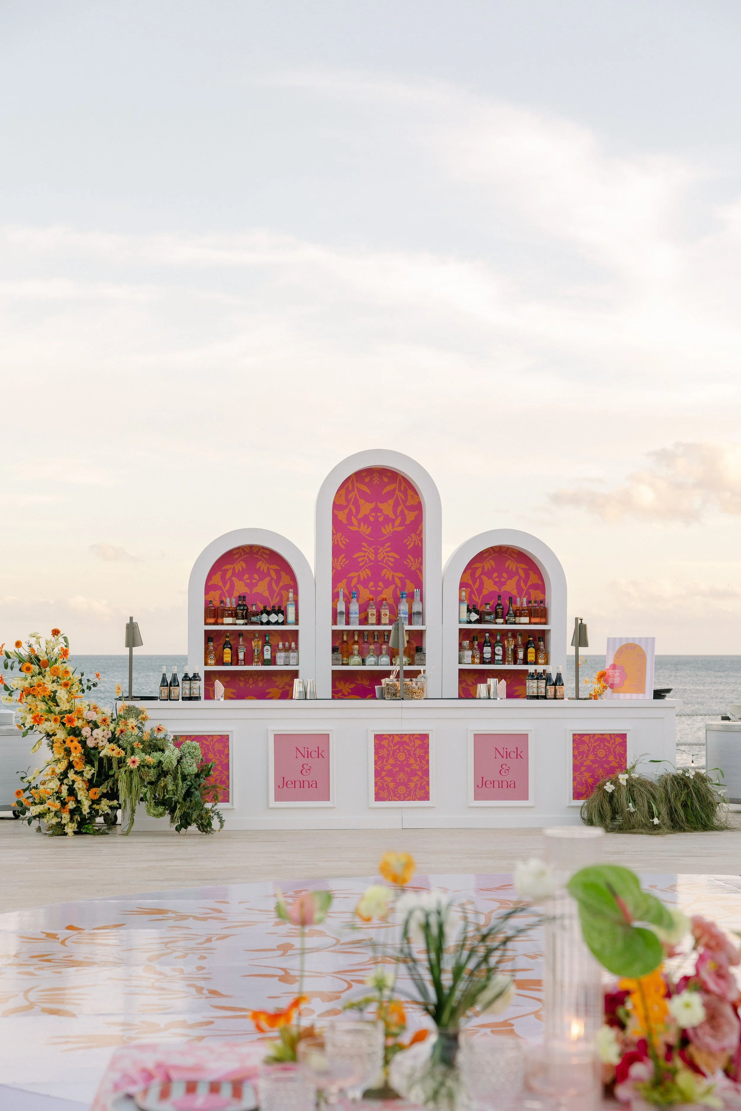 A pink and white bar setup with floral decorations, pink signs that say Nick & Jenna, and an ocean background under a partly cloudy sky, possibly at a beach wedding.