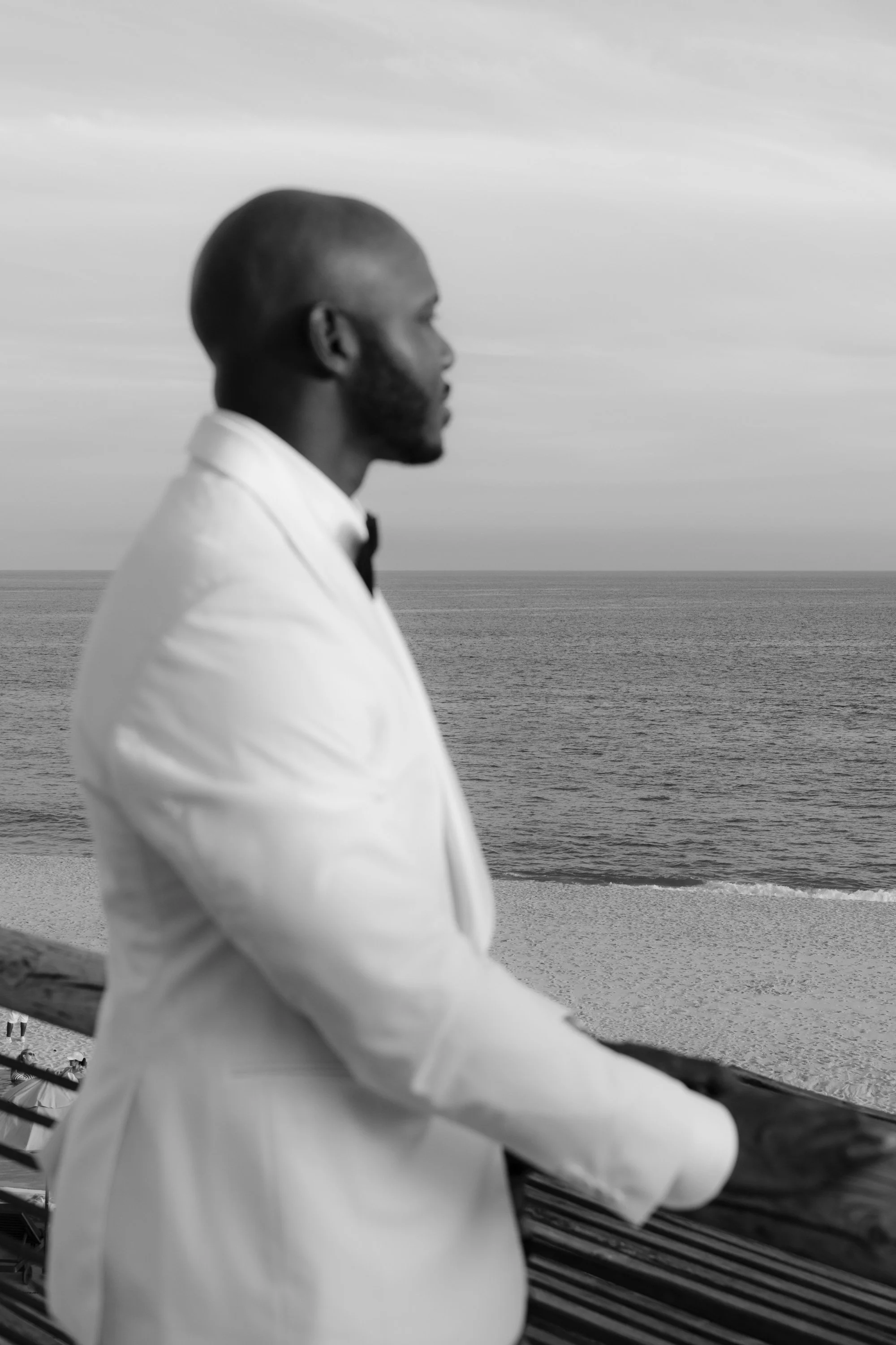A man dressed in a tuxedo with a bow tie, sitting by the ocean and looking away from the camera.
