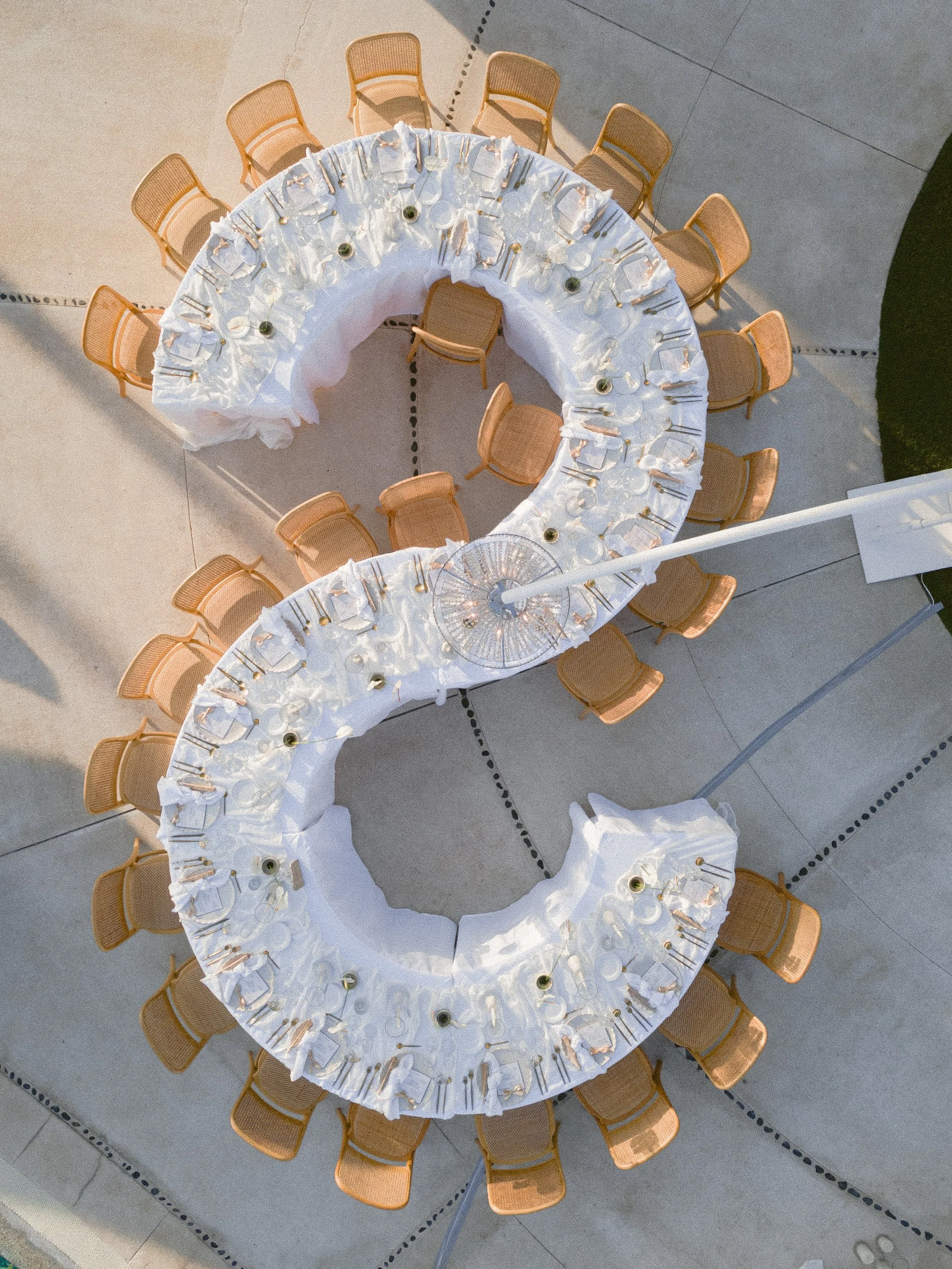 Two large round banquet tables set for an event, each covered with white tablecloths, surrounded by tan woven chairs, with place settings including white plates, napkins, and silverware, and small centerpieces.