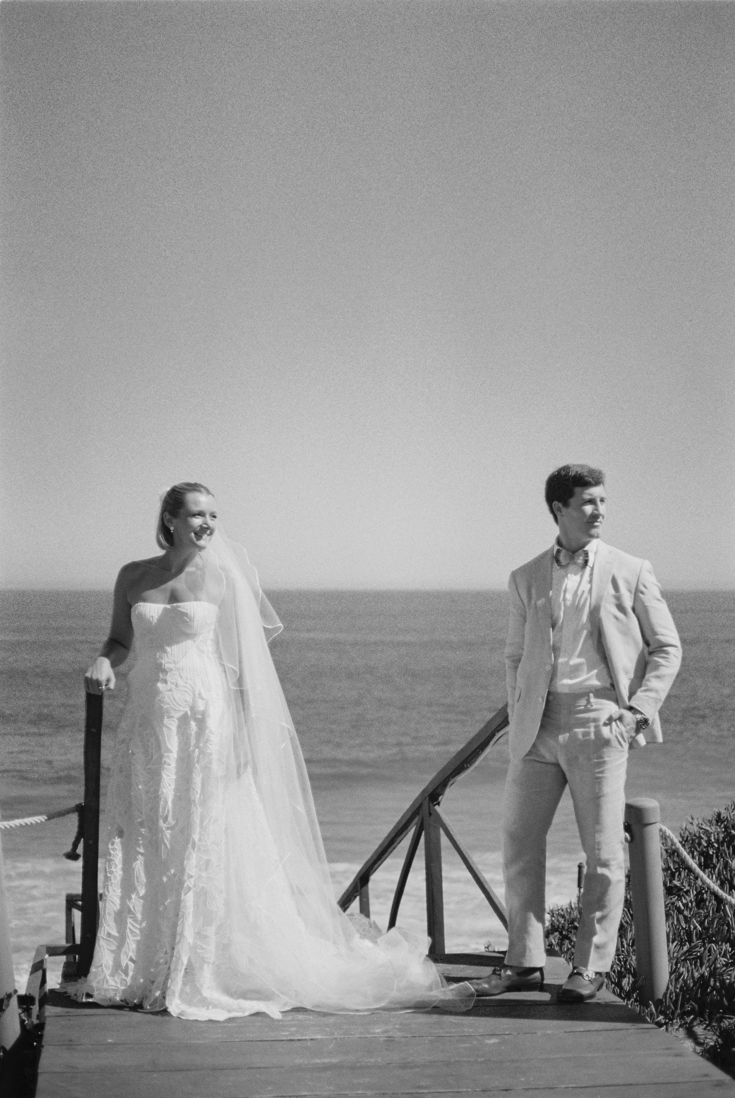 A bride and groom stand on a wooden platform outdoors, with the ocean and clear sky in the background. The bride wears a strapless wedding dress with a long train, and the groom wears a light-colored suit with a bow tie. They are smiling and looking at each other.