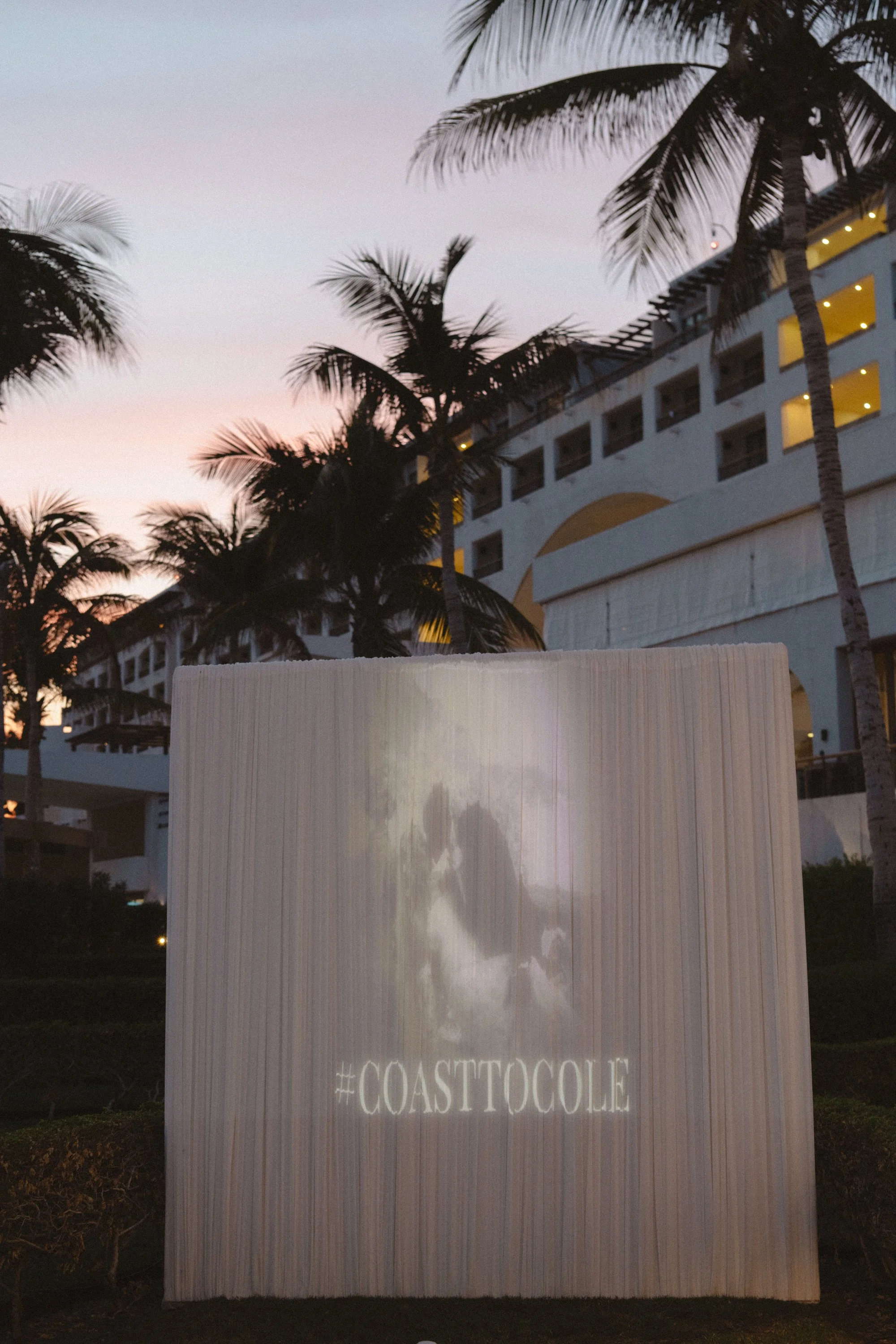 Photograph of a lit-up sign with the hashtag #COASTTOGOLE at night, with palm trees and a building with yellow-lit balconies in the background.