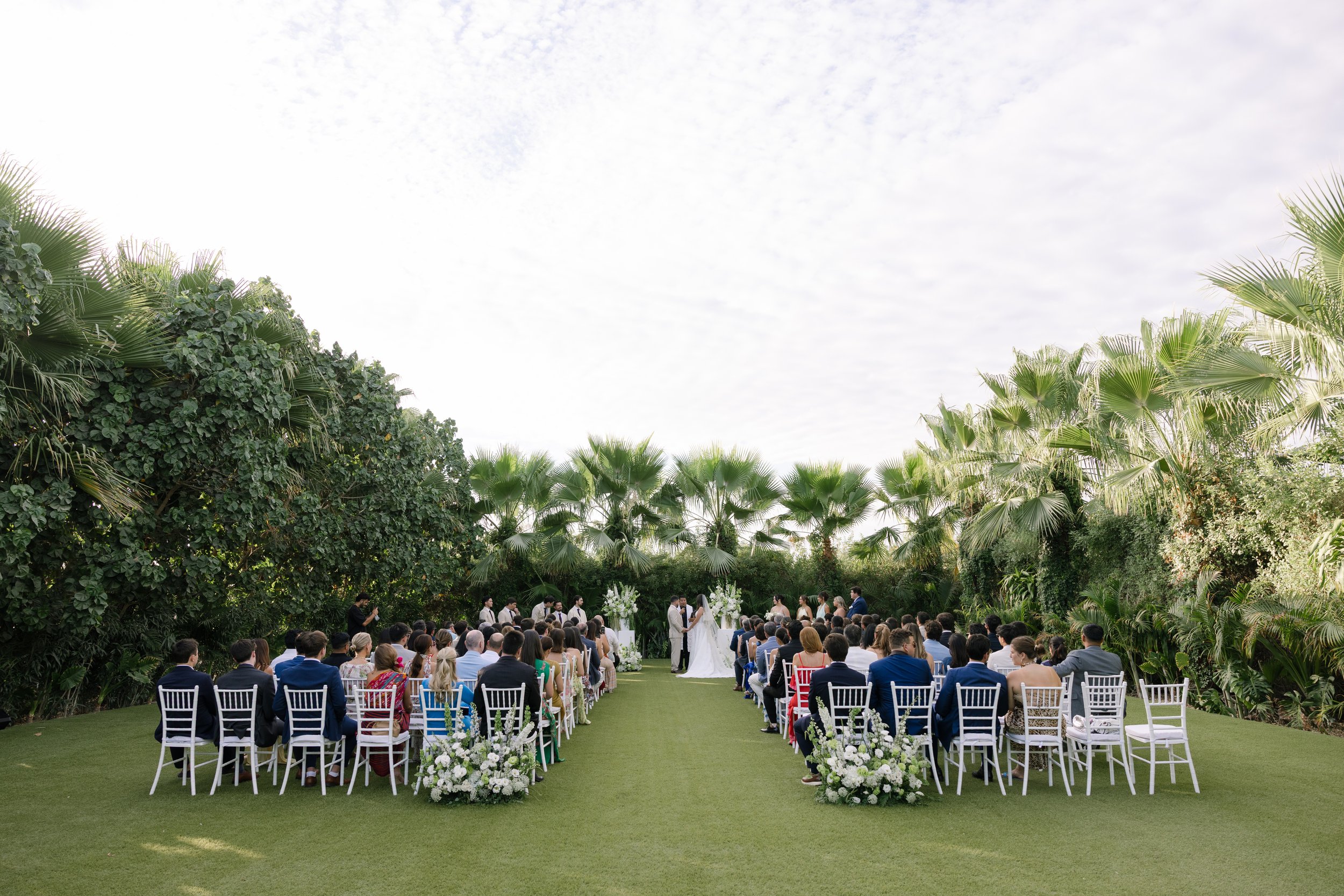 Outdoor wedding ceremony with guests seated on white chairs, floral arrangements, and a couple standing at the altar under a natural green canopy.