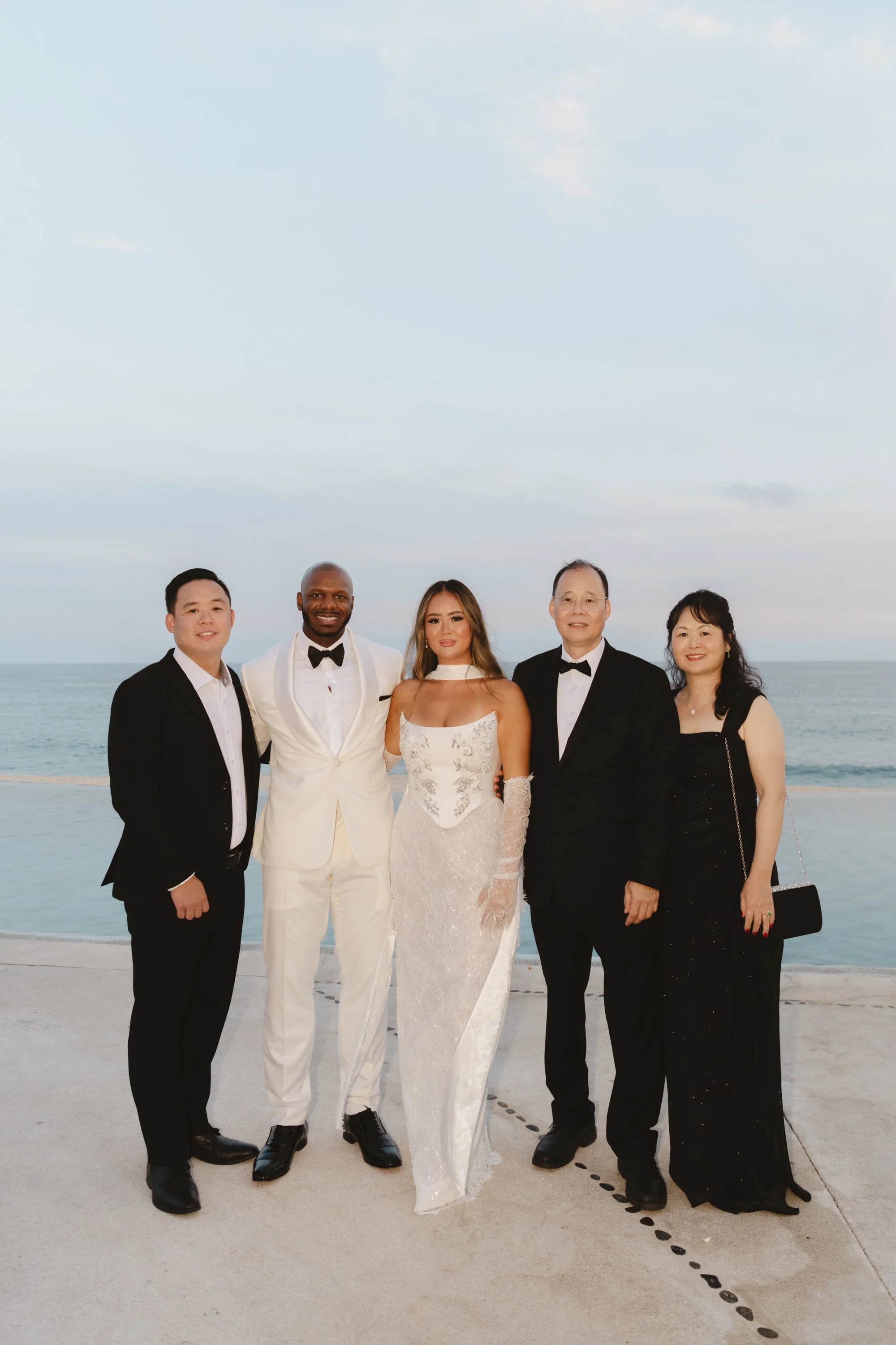 Group of five people dressed in formal attire standing outdoors near a body of water, with a cloudy sky overhead.