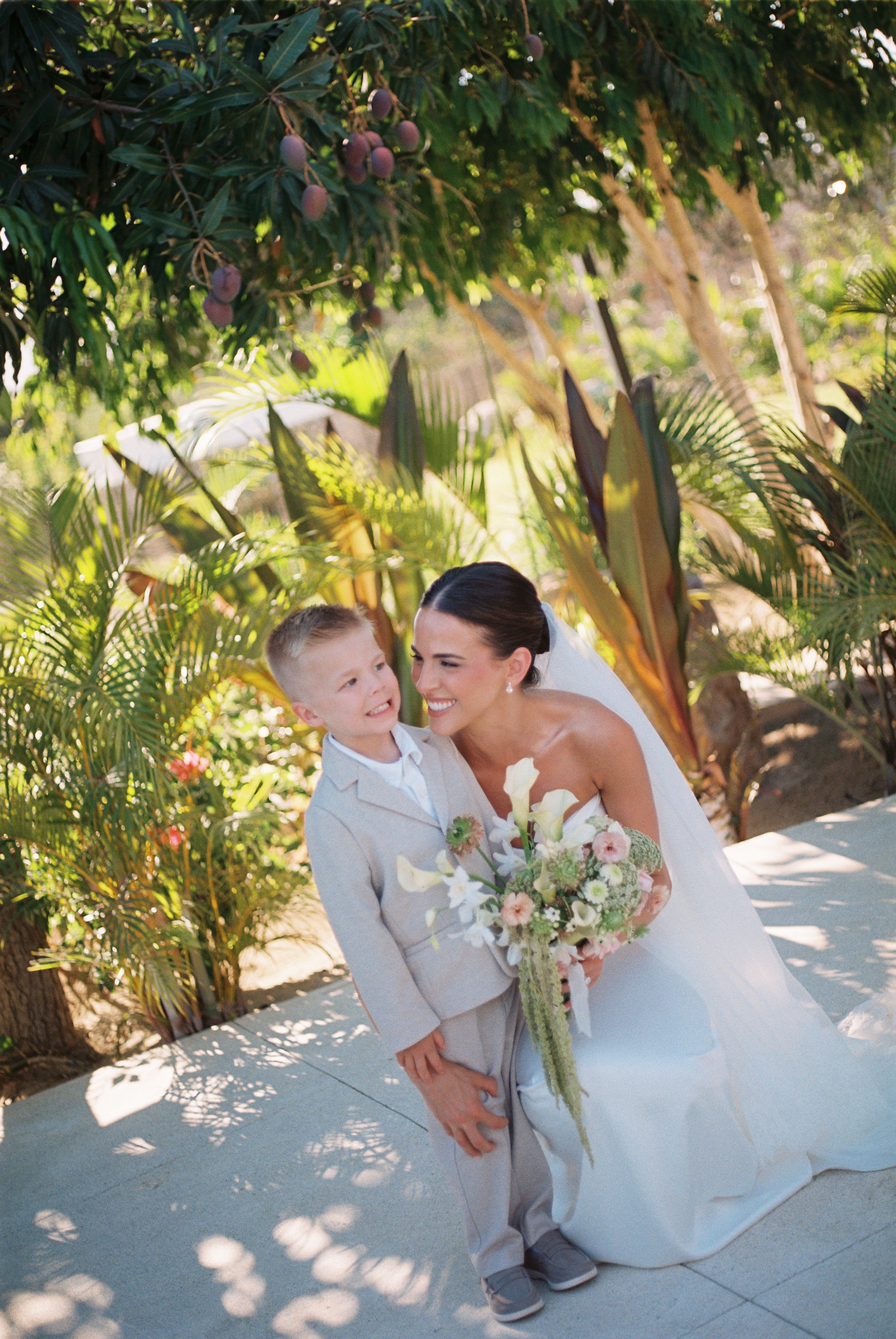 A bride in a white wedding dress holding a bouquet, smiling at a young boy in a gray suit and tie, outdoors with lush green plants and trees in the background.