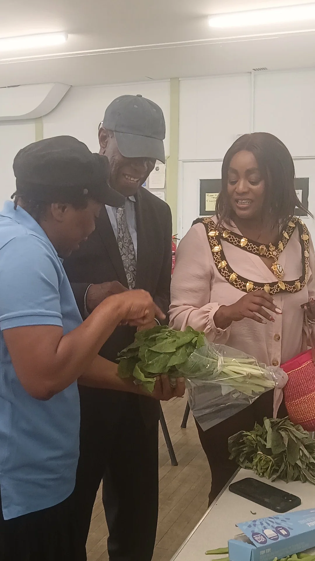 Three people in a room looking at a bunch of leafy greens, with one person holding a bouquet of greens and another holding a basket, with a table in front.