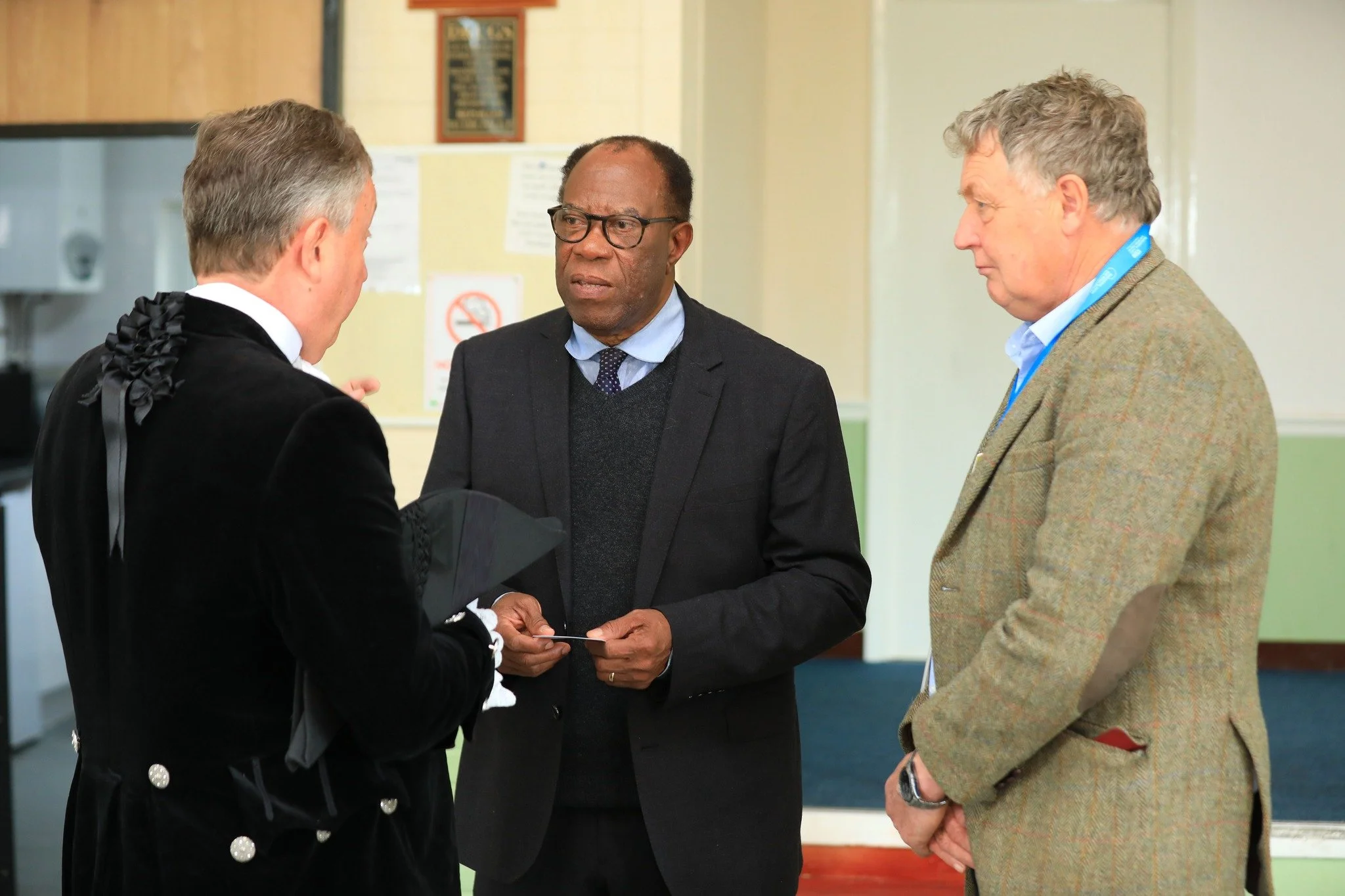 Three men engaged in conversation in an indoor setting, one wearing a judge's robe, the second in a suit with glasses, and the third in a blazer with a blue lanyard. The background includes a no-smoking sign and a chalkboard.