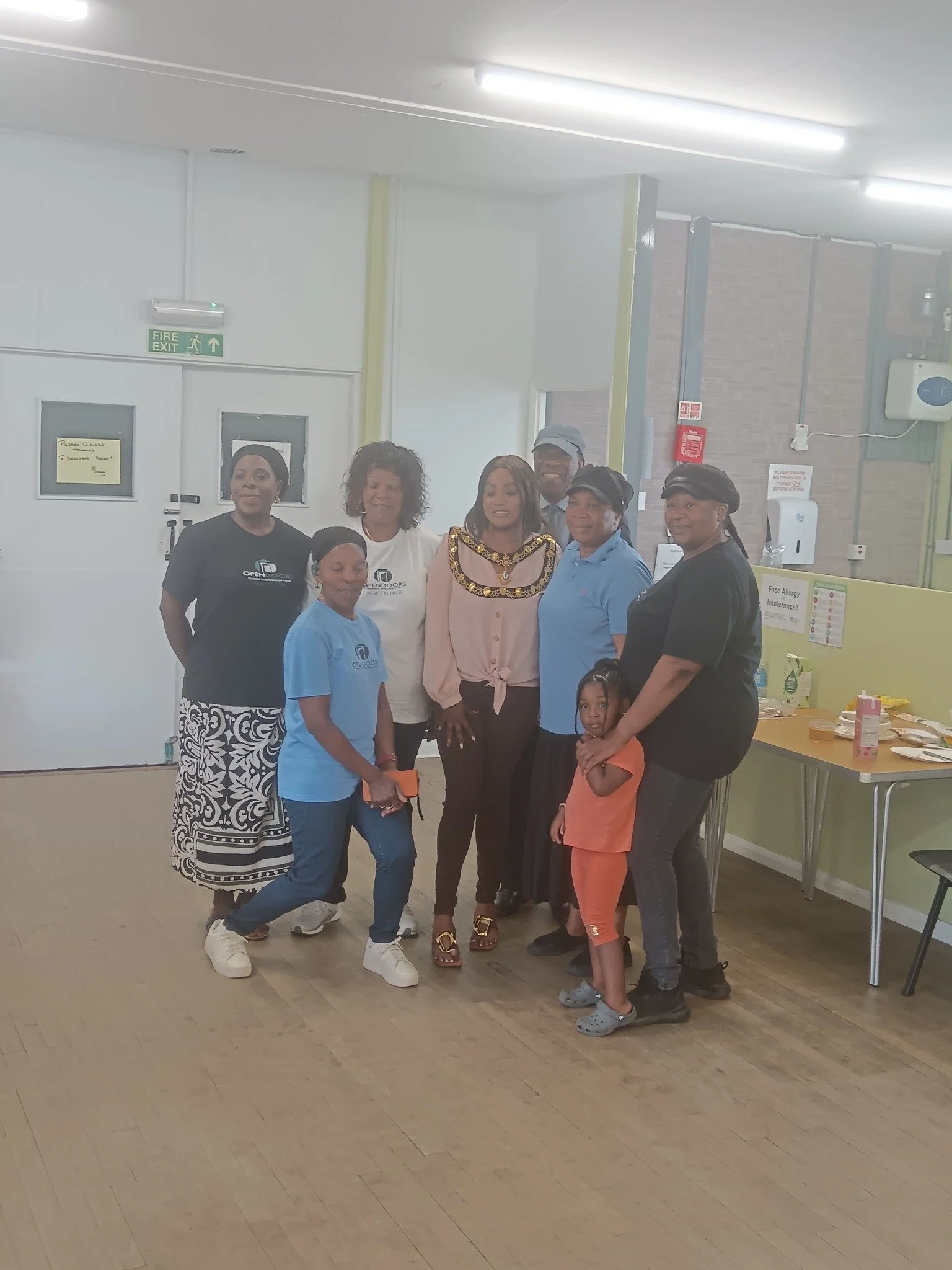 A group of eight people, including children, women, and men, are standing together in an indoor community or event space, smiling at the camera.