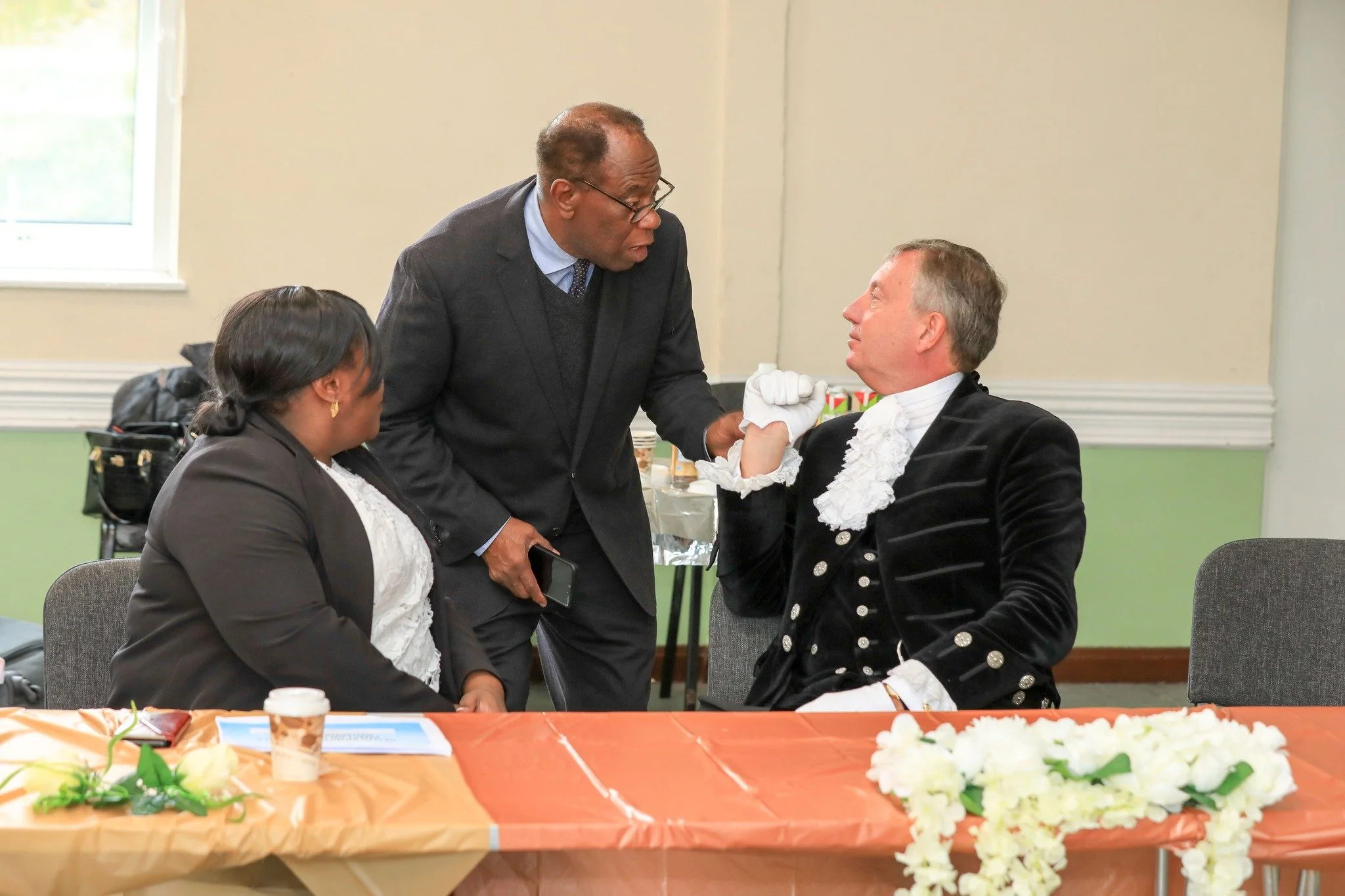 Three people engaged in conversation at a formal event, sitting around a table with a floral centerpiece, with a man wearing a dark suit and glasses standing and two seated individuals, one in a dark blazer and the other in a tuxedo with a ruffled shirt.