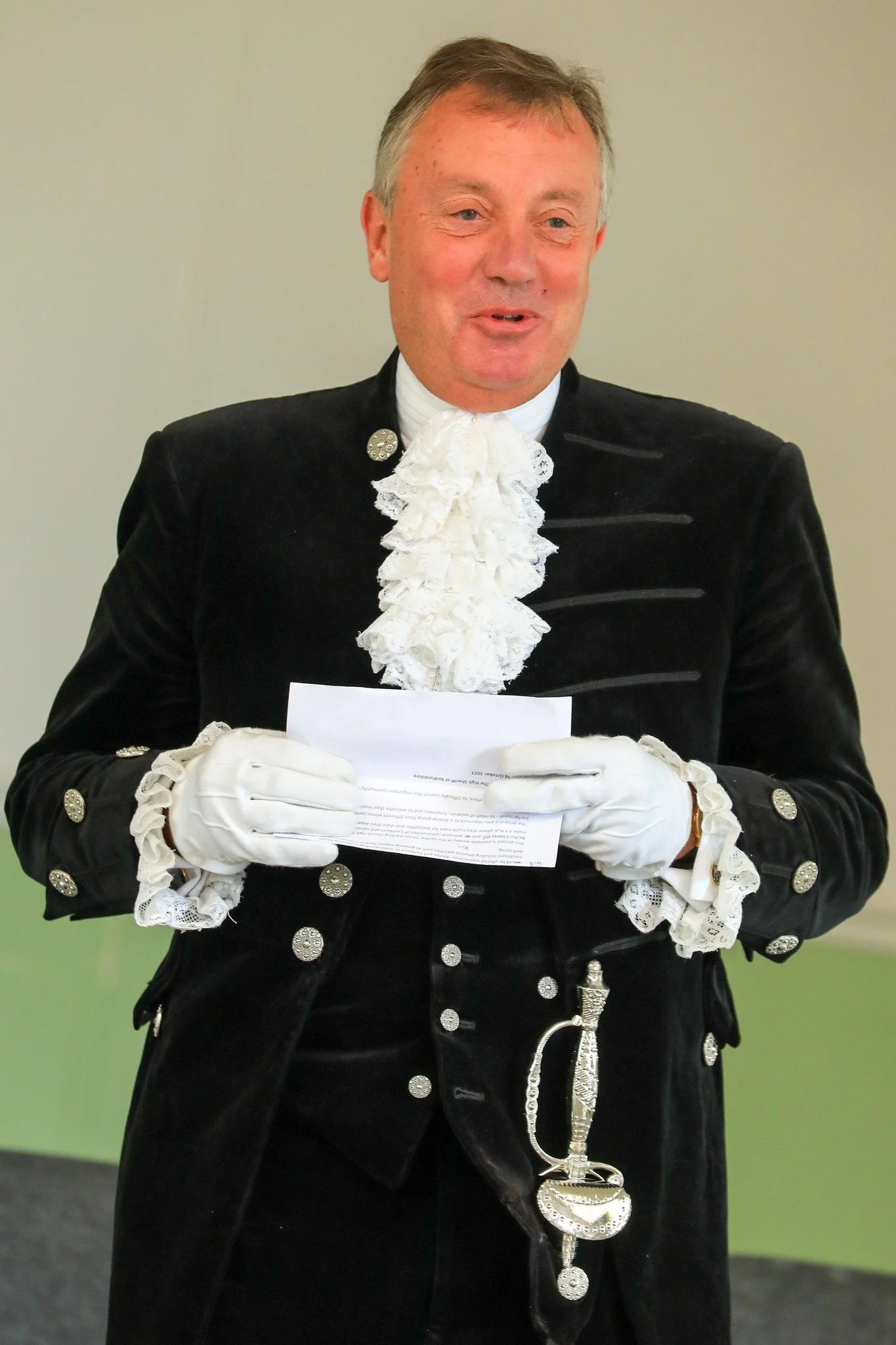 A man dressed in a traditional formal black jacket with silver buttons, a white lace cravat, and white gloves, holding a piece of paper.