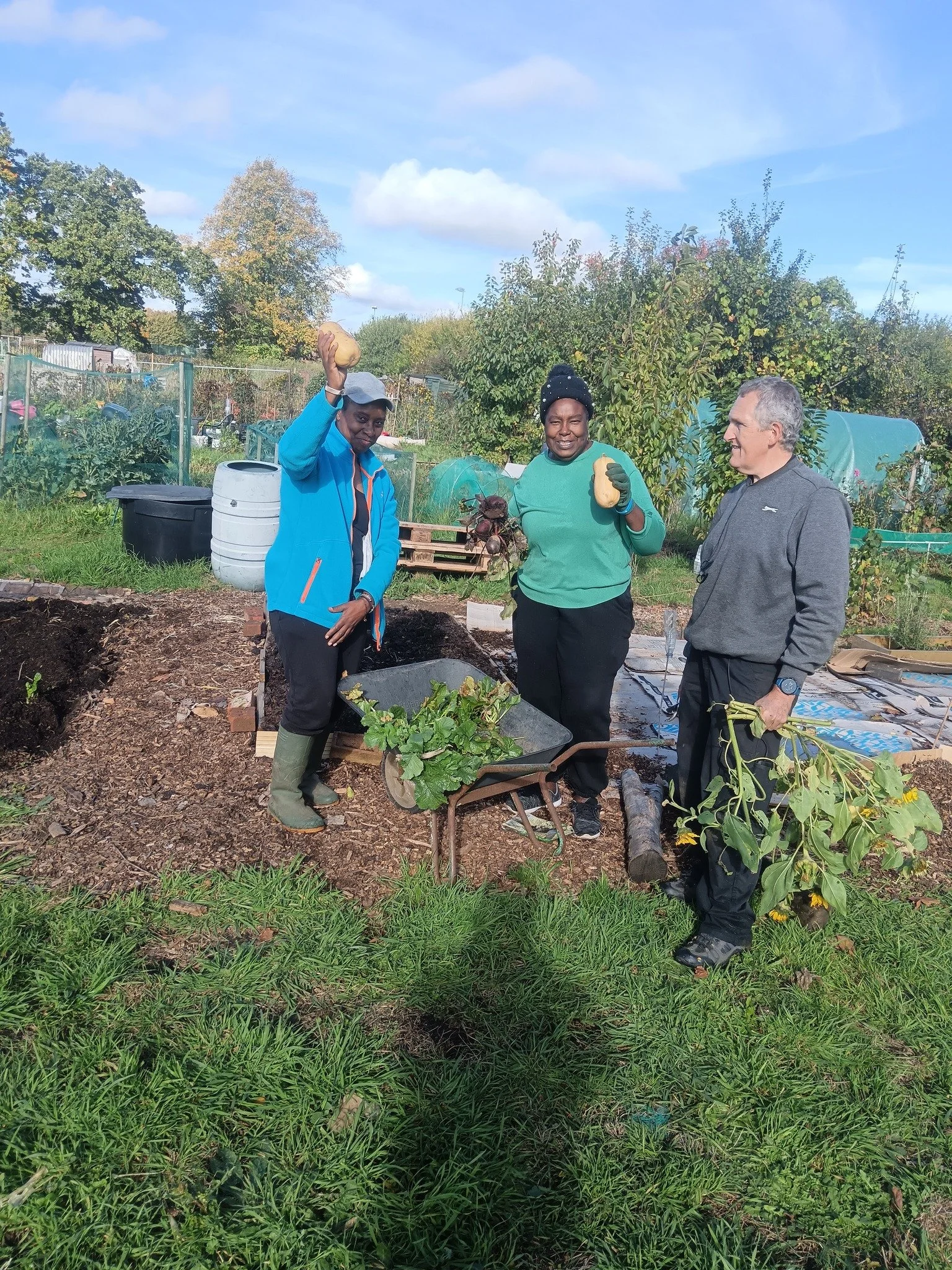 Three people standing in a garden, two women and one man, holding vegetables and flowers, with trees and garden beds in the background on a sunny day.